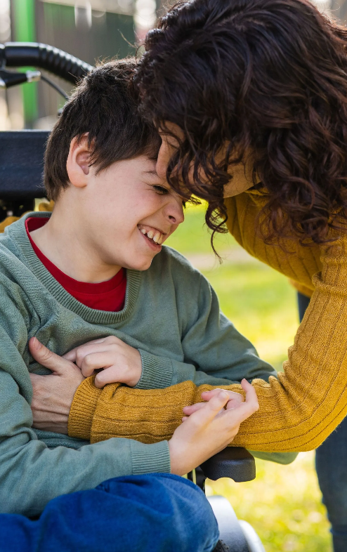 A young boy smiling and laughing while pressing his head against a woman who is leaning over him outdoors.