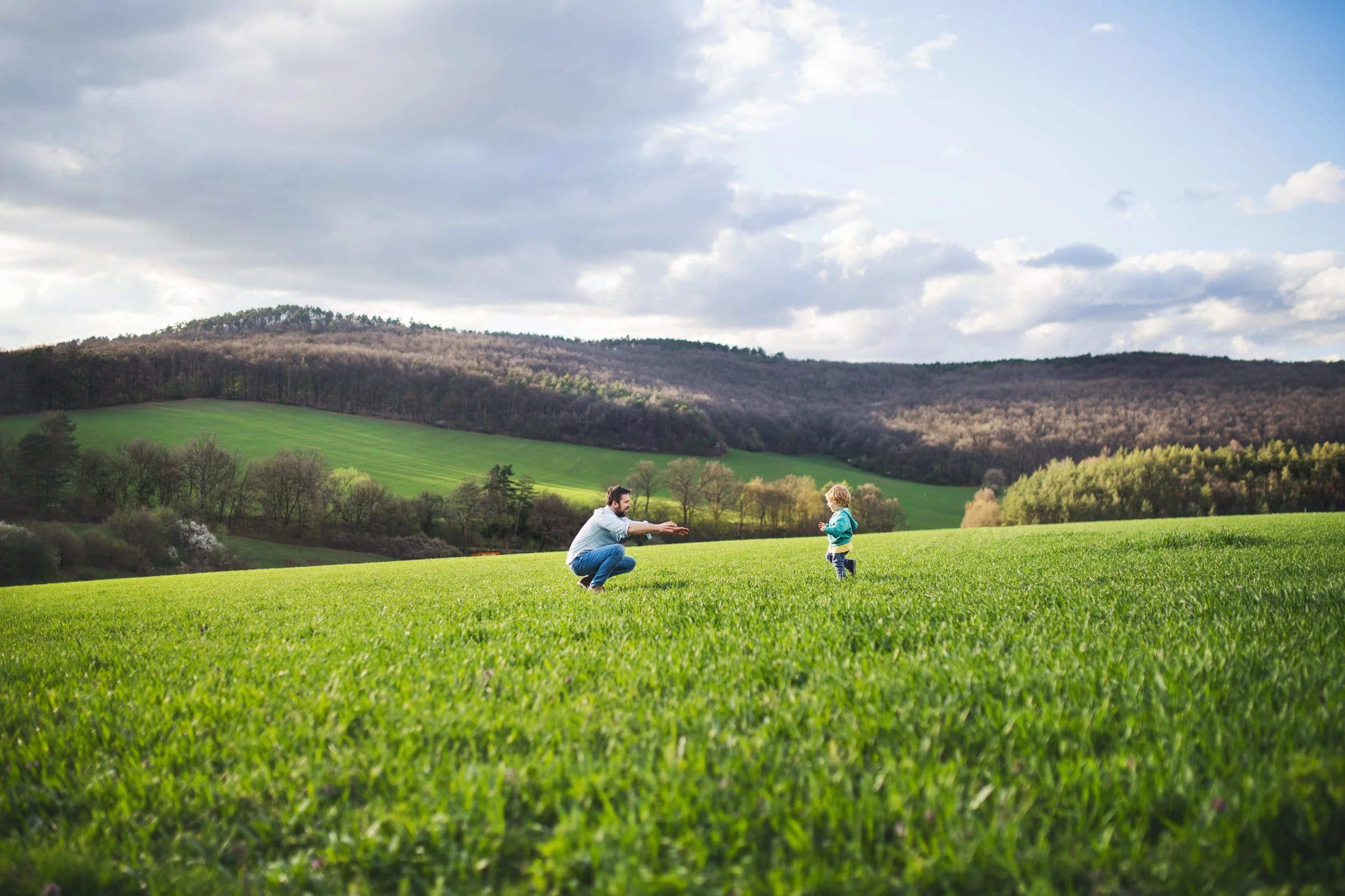 A man and a young child playing outdoors on a vast green grassy field with rolling hills and trees in the background under partly cloudy skies.