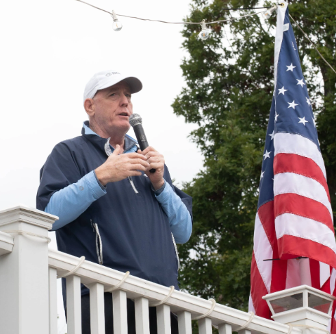 An older man speaking into a microphone on a porch or balcony, with an American flag beside him and trees in the background.