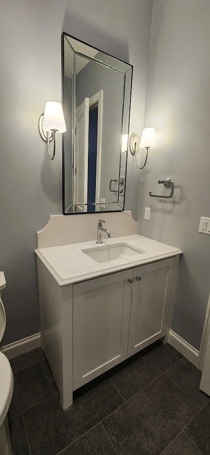 Bathroom vanity with a white cabinet, a sink, a large mirror, two wall sconces, and towel holder on a gray wall.