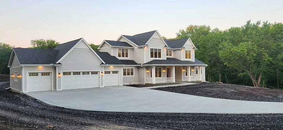 Newly constructed two-story white house with three garage doors and a spacious concrete driveway, surrounded by trees.