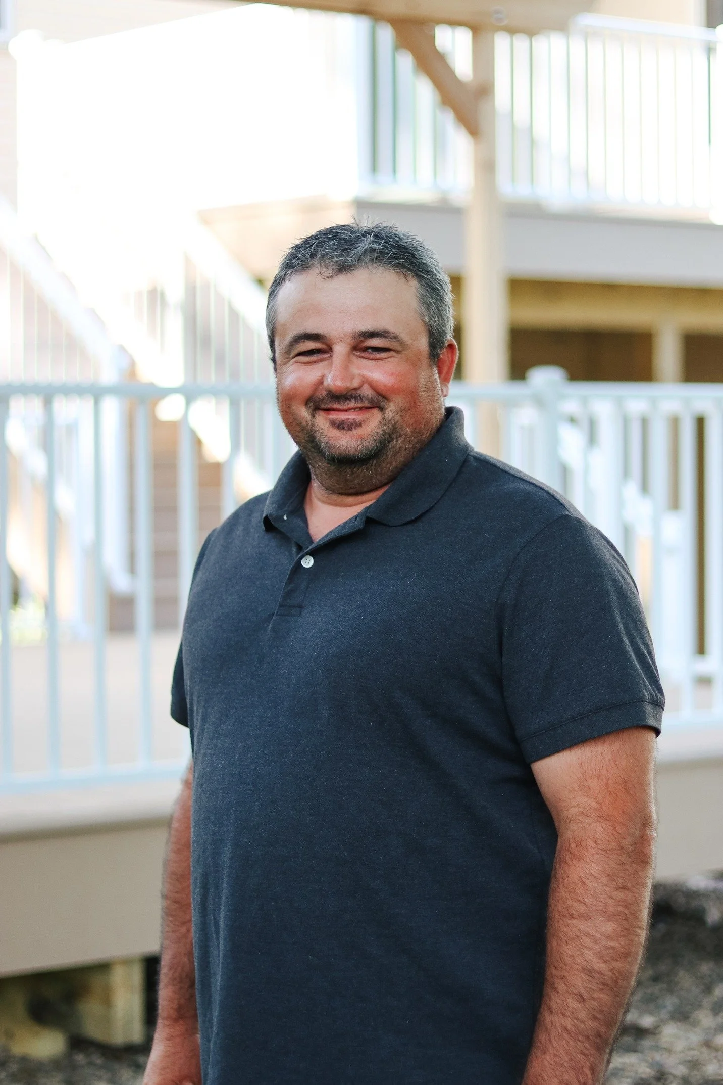 A man with short, dark hair and a beard standing outdoors, smiling, next to a white fence and a wooden staircase, with sunlight coming through the railing.