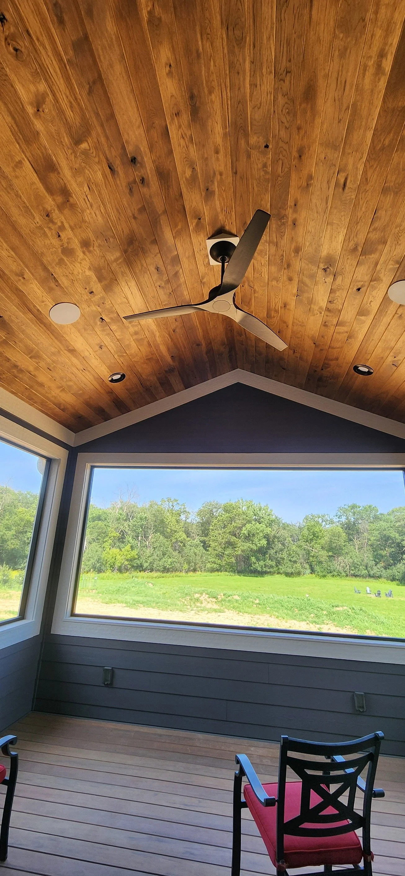 A covered porch with a wooden ceiling, ceiling fan, and large windows overlooking a green landscape with trees.