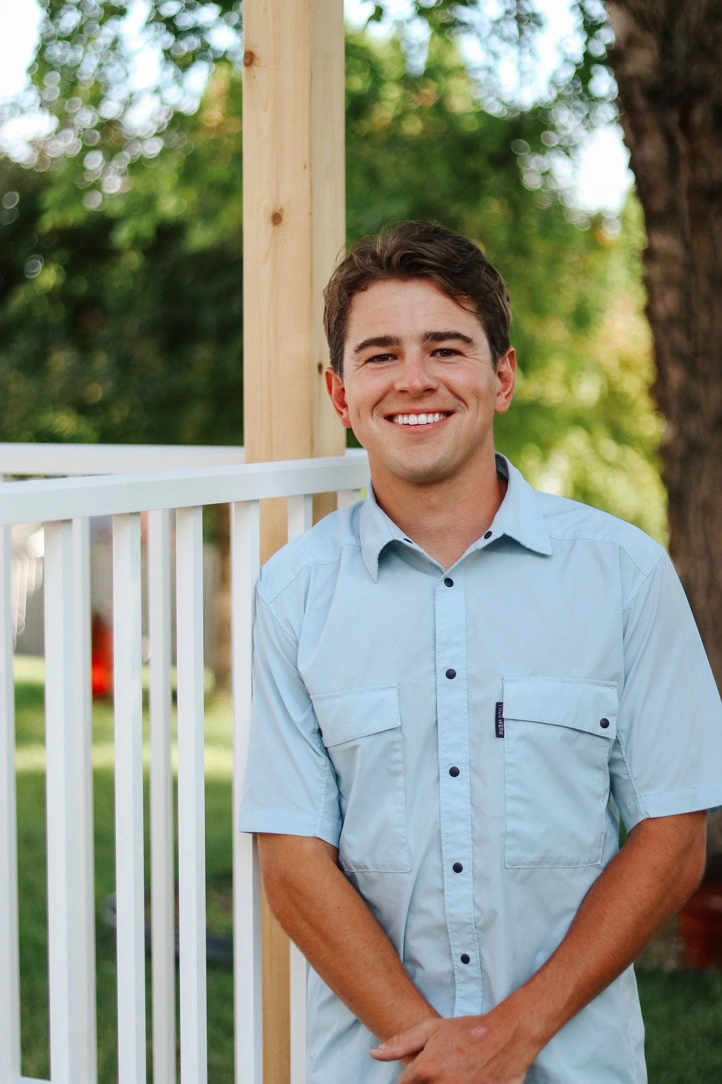 A young man with dark hair and a light blue shirt smiling outdoors, standing near a white fence and a wooden post, with green trees and a blurred background.