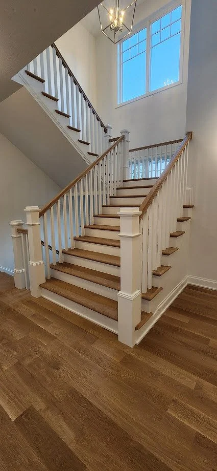 A staircase in a modern home with white risers, wooden treads, and a white railing with wooden handrails, leading to an upper floor with large windows and a chandelier hanging from the ceiling.