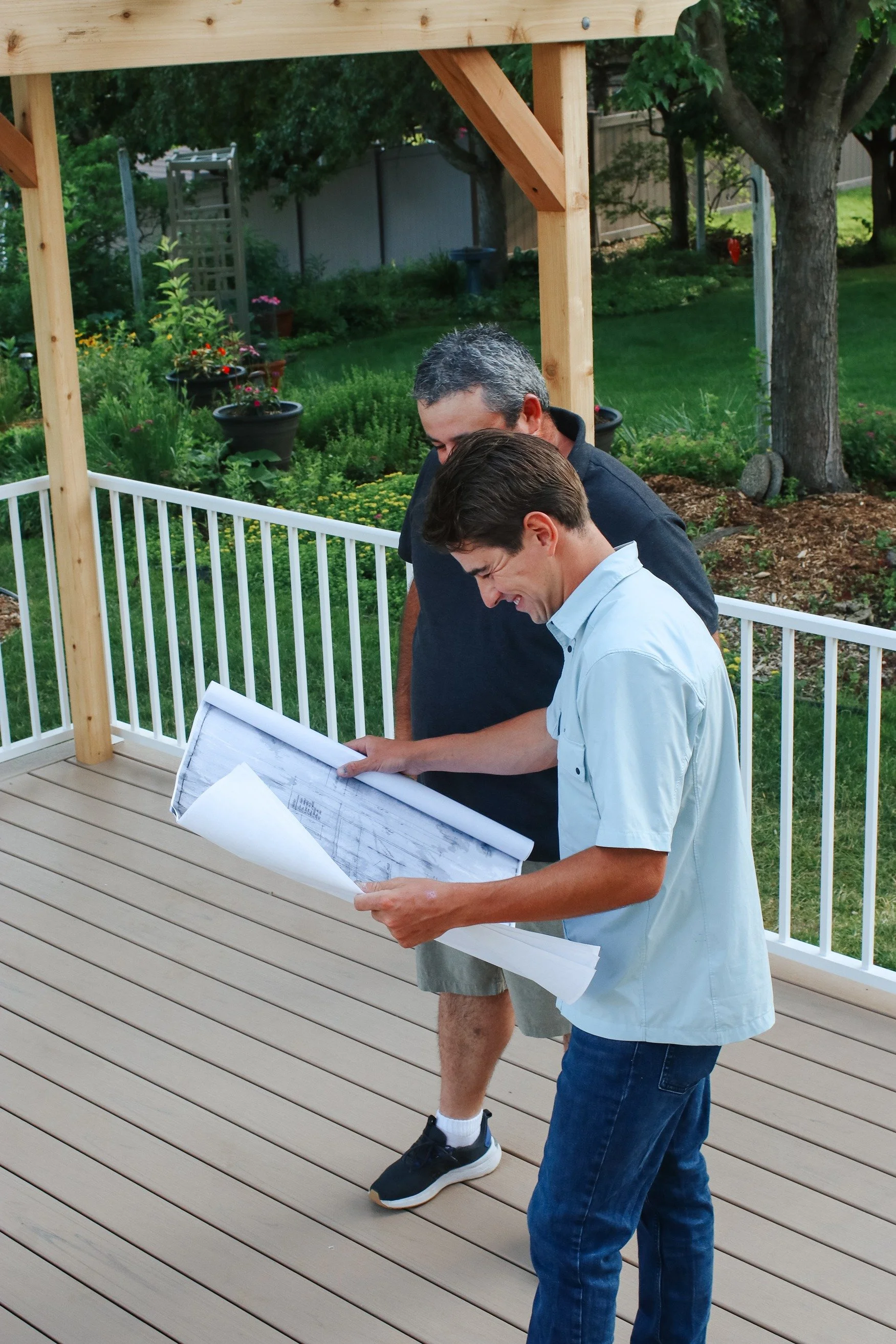 Two men looking at blueprints on a balcony under construction, with a wooden deck and green backyard in the background.