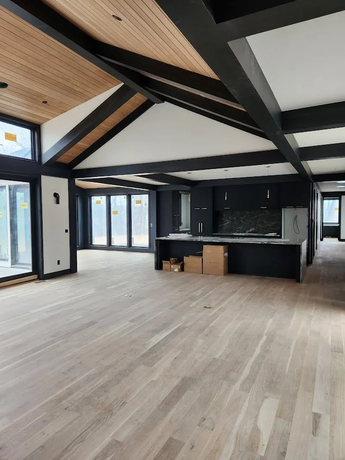 Empty modern open-concept living space with large windows, black kitchen cabinets, marble backsplash, and light wood flooring, with visible ceiling beams and some cardboard boxes on the floor.