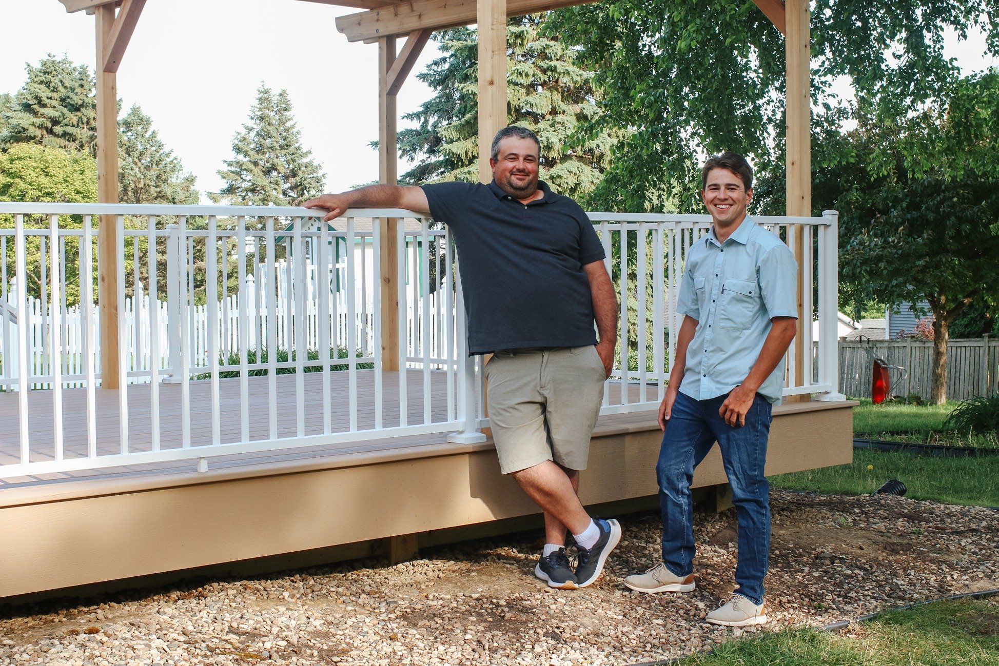 Two men standing on a deck in a backyard, smiling at the camera. One is leaning on a white railing, the other standing nearby. The deck has a wooden frame and is surrounded by trees and a white picket fence.