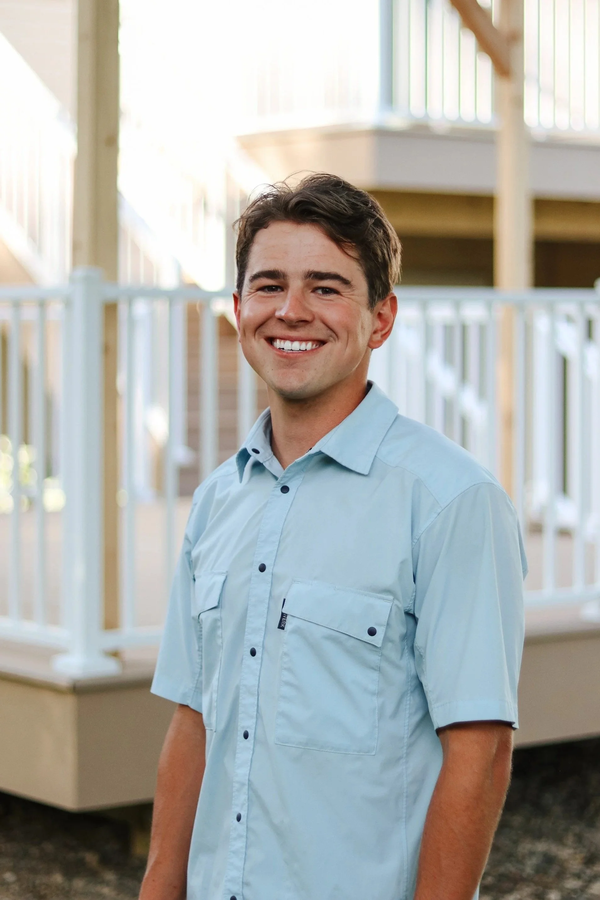 A young man with brown hair and a bright smile standing indoors near a staircase with wooden railings, wearing a light blue button-up shirt.