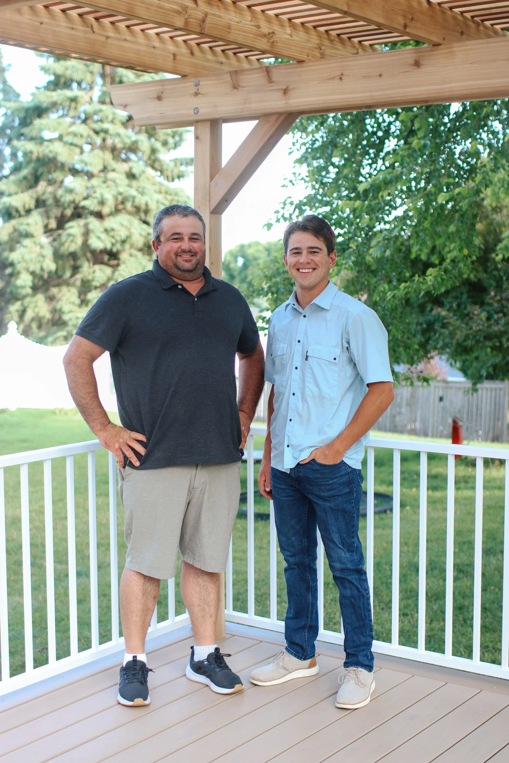 Two men standing on a wooden deck with a white railing, outdoors near trees, smiling at the camera.