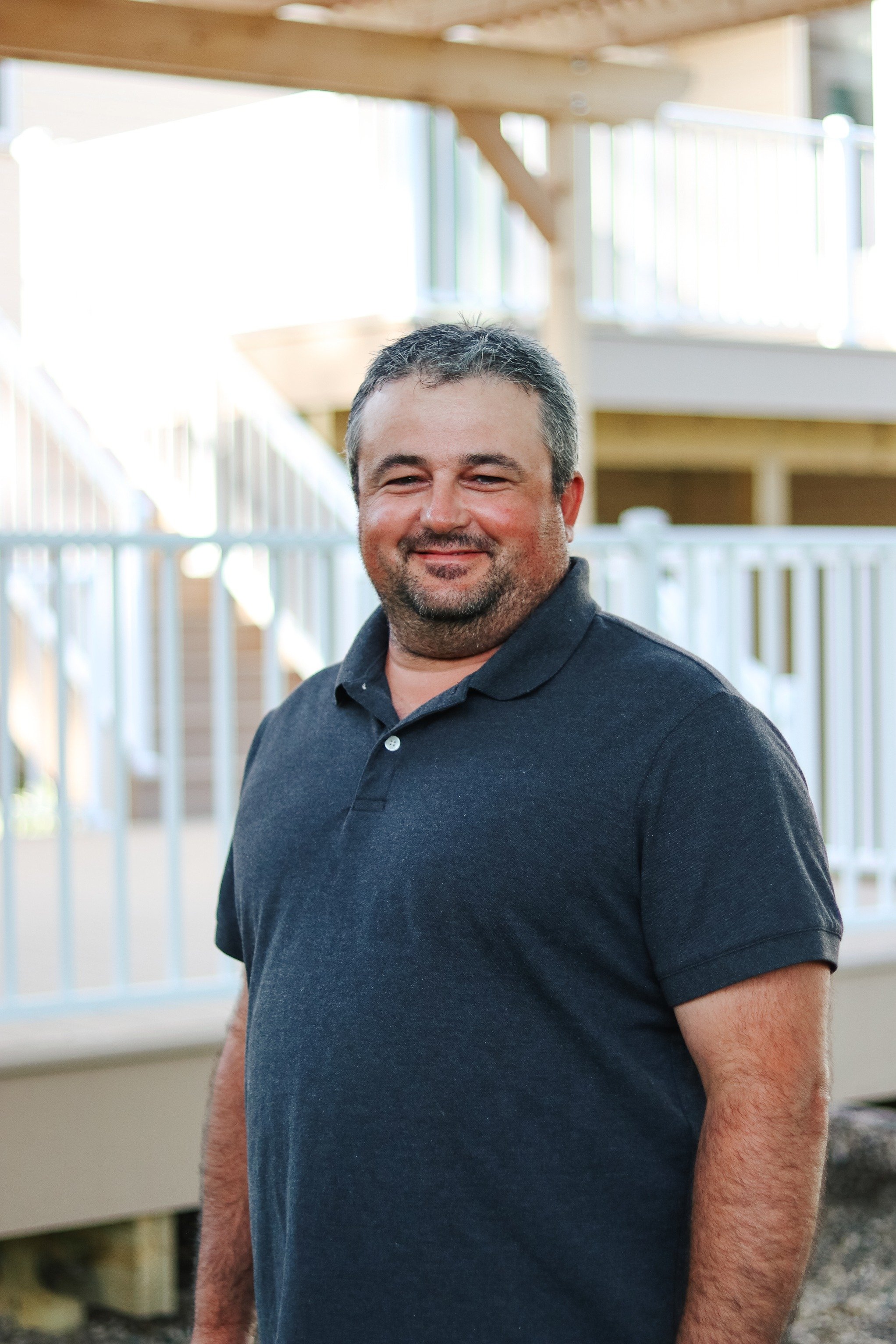 Portrait of a smiling man with short dark hair and facial hair, wearing a black polo shirt, standing outdoors near a wooden staircase and a white picket fence, bright and sunny day.