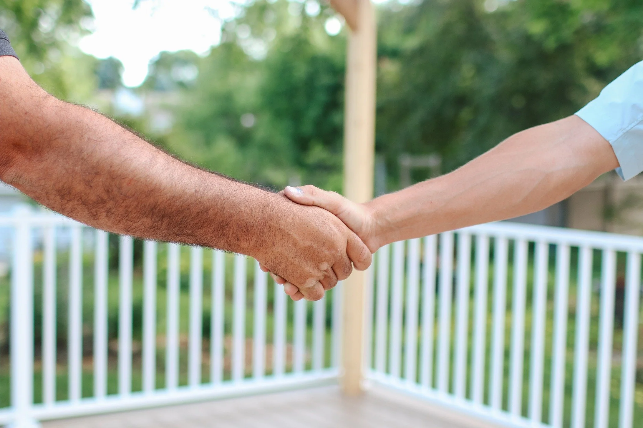 Two people shaking hands outdoors with a white fence and green trees in the background.