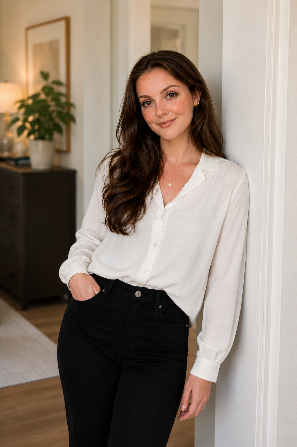 A young woman with long wavy brown hair leaning against a white wall, wearing a cream-colored blouse and black pants, in a cozy room with a dresser, a potted plant, and framed artwork in the background.
