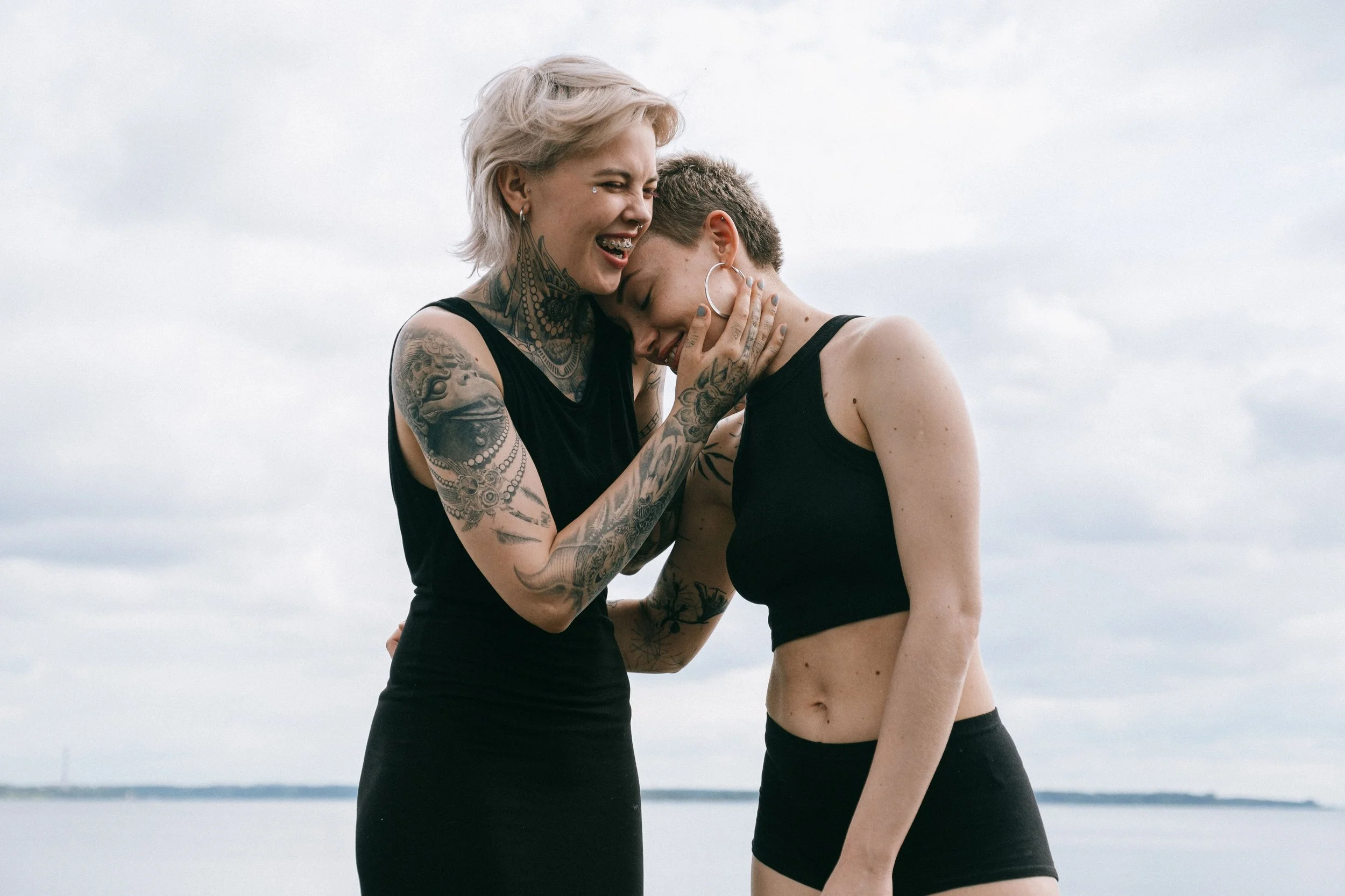 Two women with short hair and tattoos embracing each other. They are wearing sleeveless black tops, standing outdoors, with a cloudy sky background.