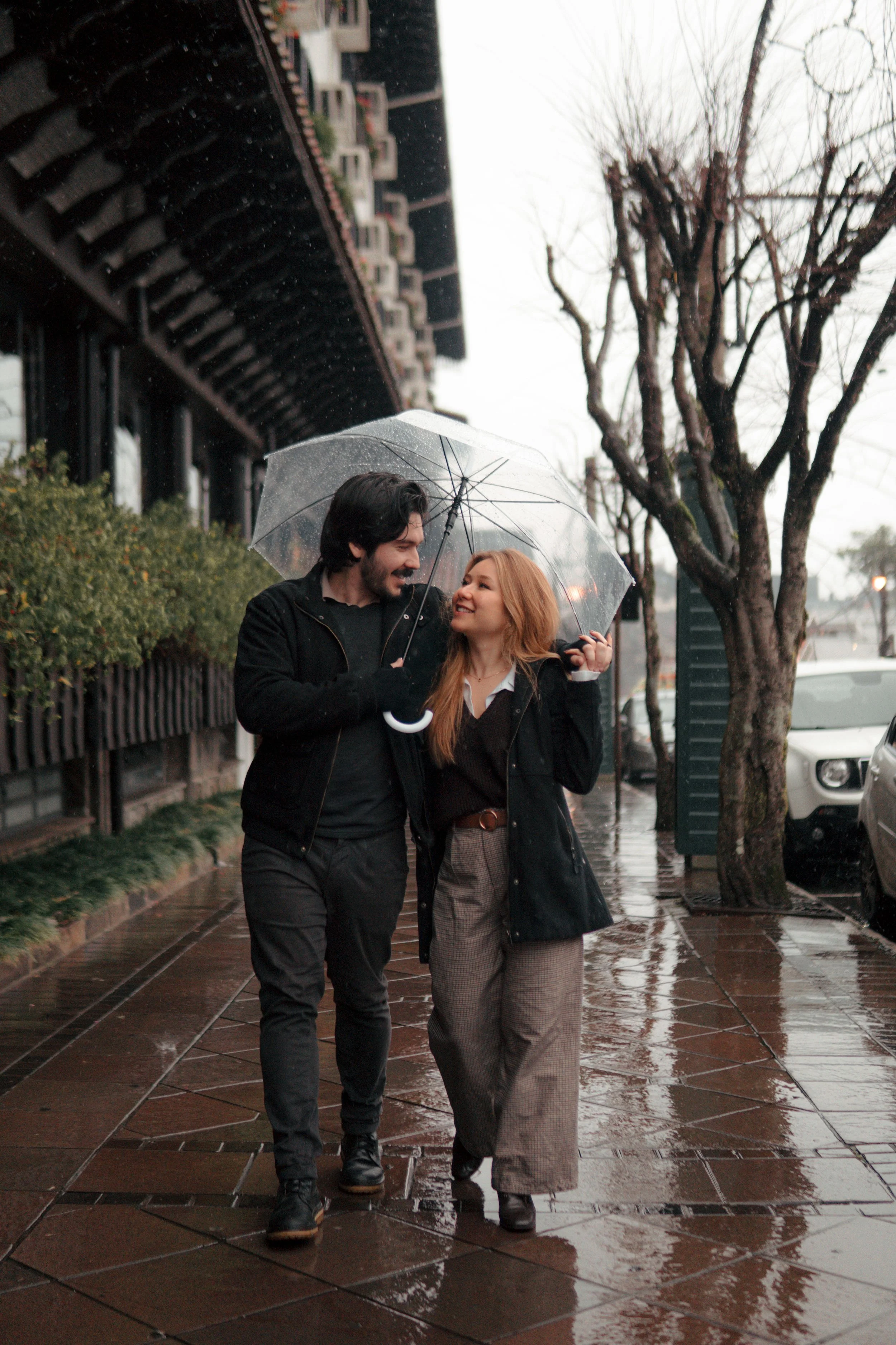 A man and woman walking together in the rain under a clear umbrella, smiling and looking at each other, with wet pavement, trees, and buildings in the background.