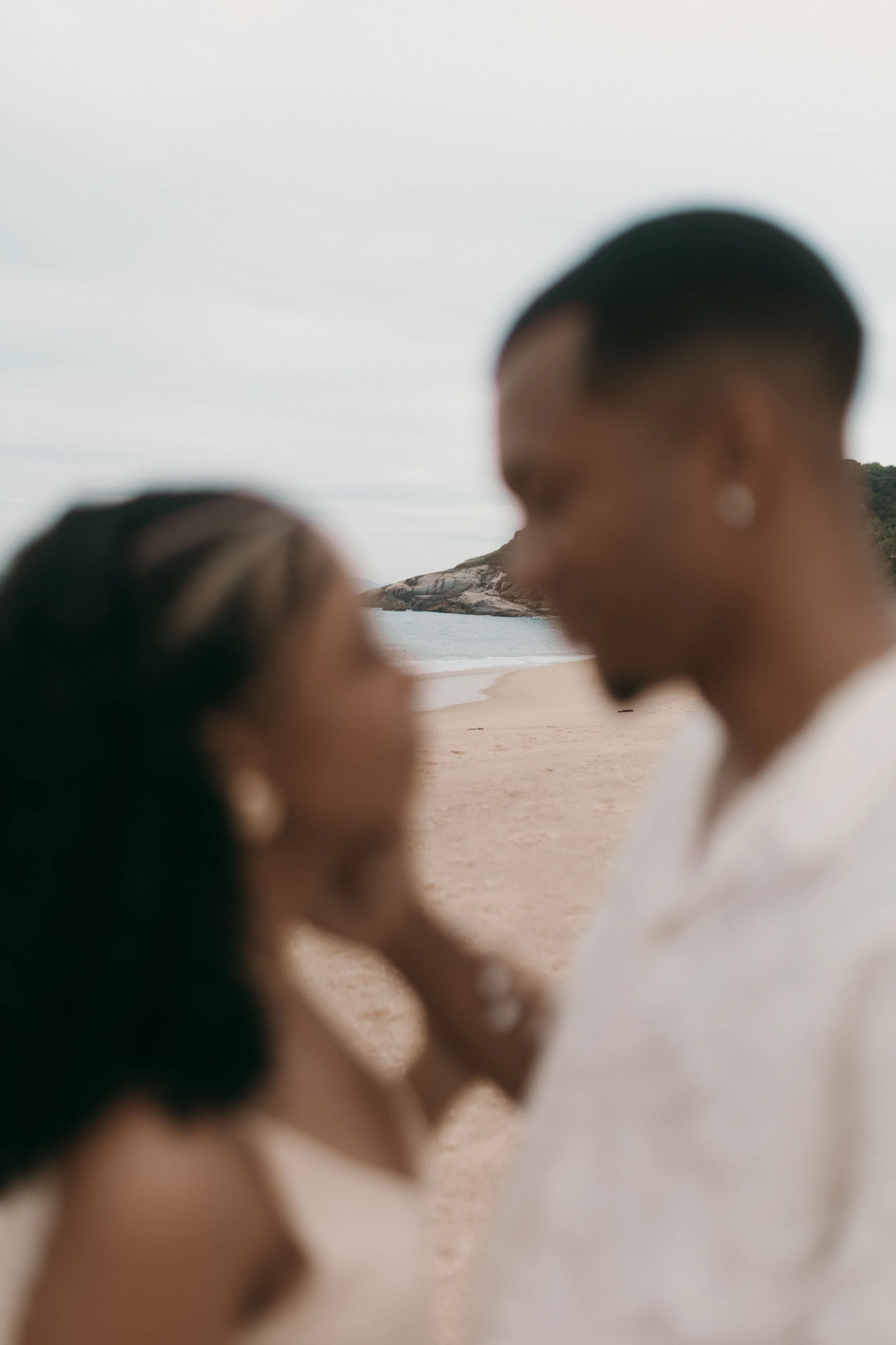 A blurred image of two people close to each other on a beach with rocks and water in the background.