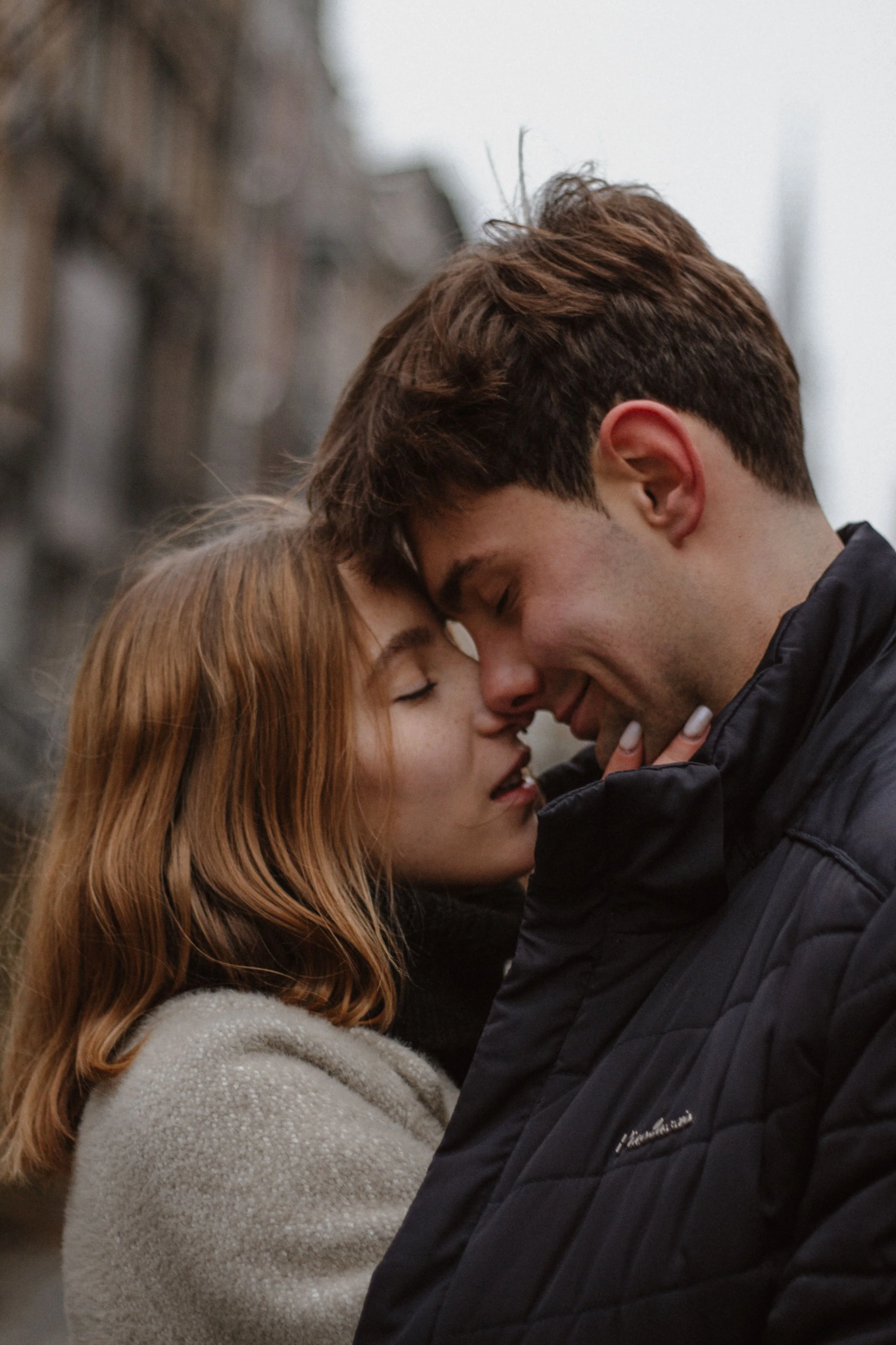 A young couple with eyes closed, touching foreheads and smiling while embracing outdoors on a cloudy day.