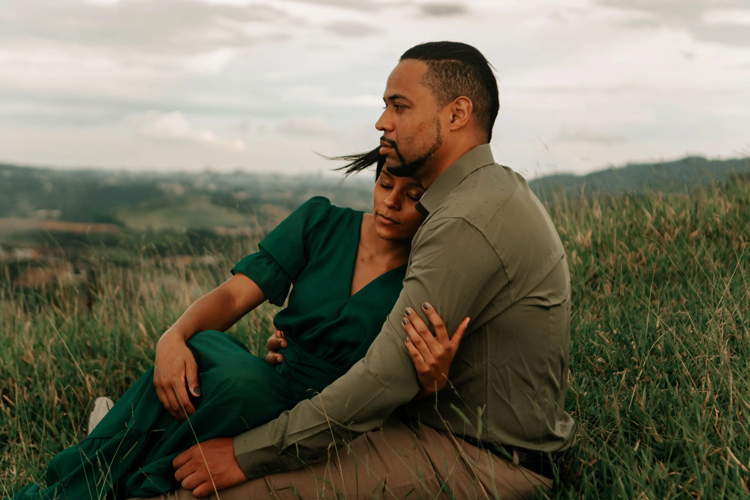 A man and woman sit together in a grassy field under a cloudy sky, embracing each other with the woman's head resting on the man's chest and his arms around her.