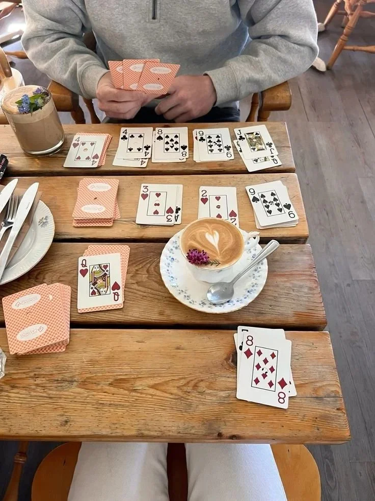 Person playing cards at a wooden table in a cafe with a latte and a flower in a cup.