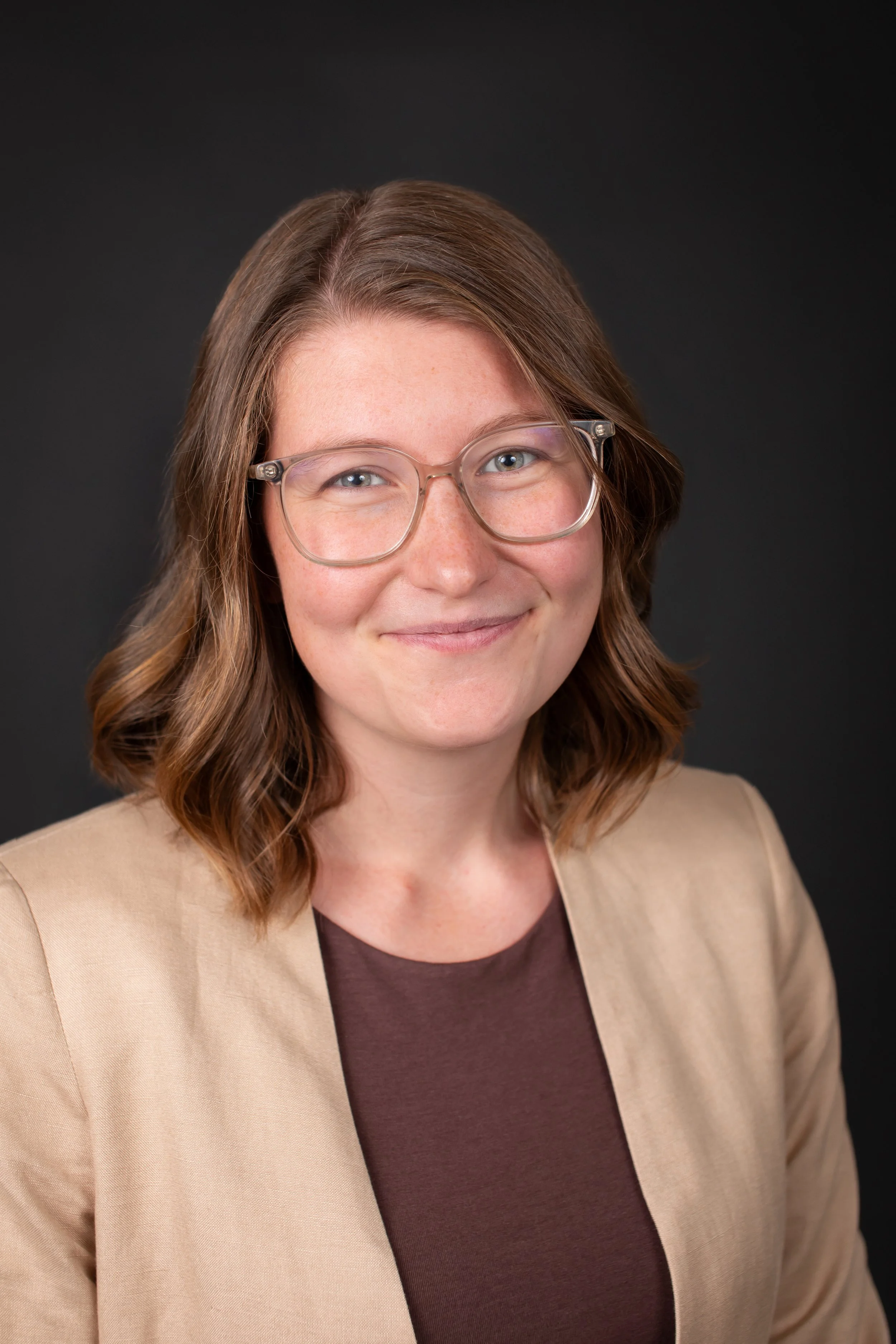Portrait of a woman with shoulder-length wavy brown hair, wearing round glasses, a beige blazer, and a brown top against a dark background.