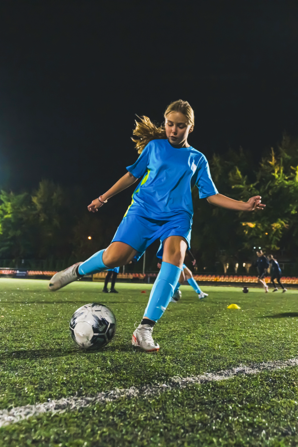 A young girl in a blue soccer uniform kicking a soccer ball on a field at night, with other players and trees in the background.