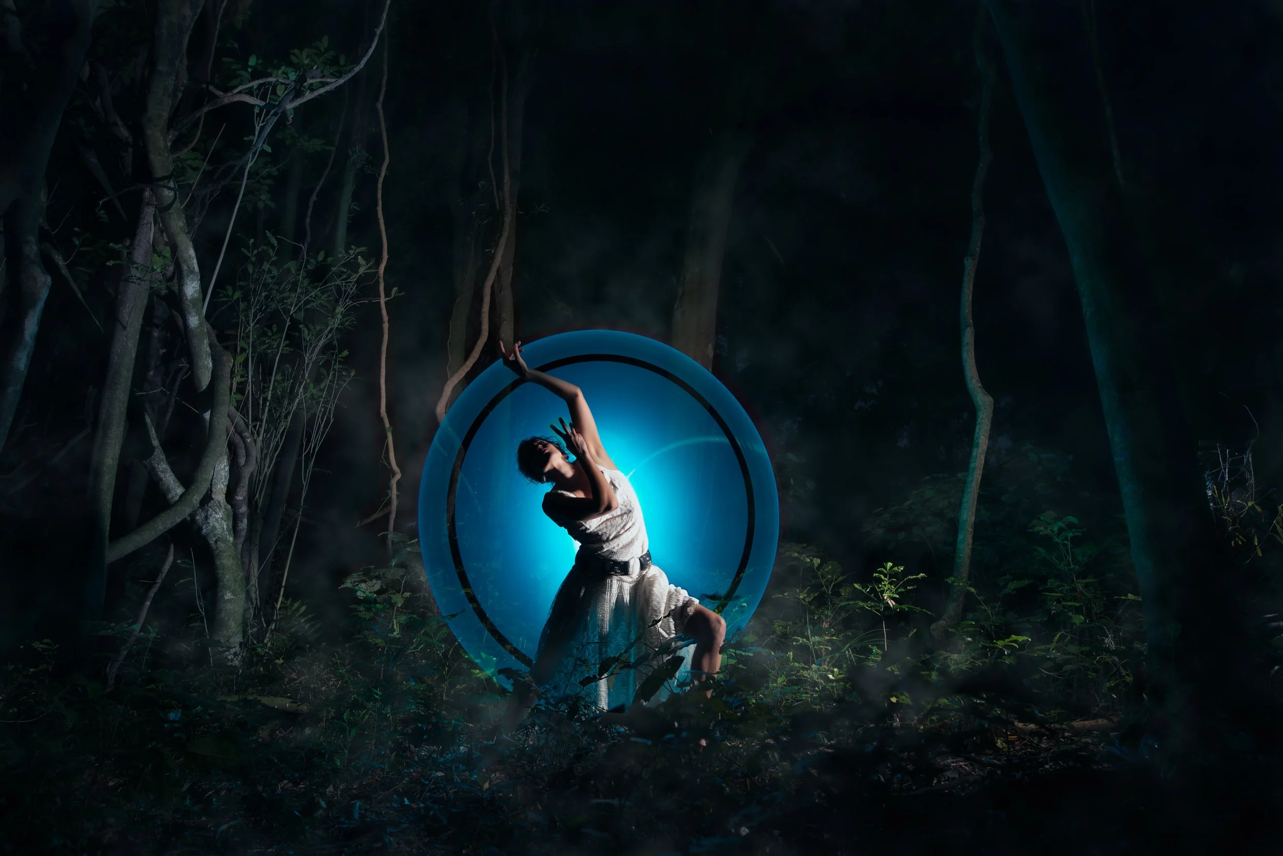 A woman in a white dress poses inside a glowing blue ring outdoors at night, surrounded by dark trees and mist.