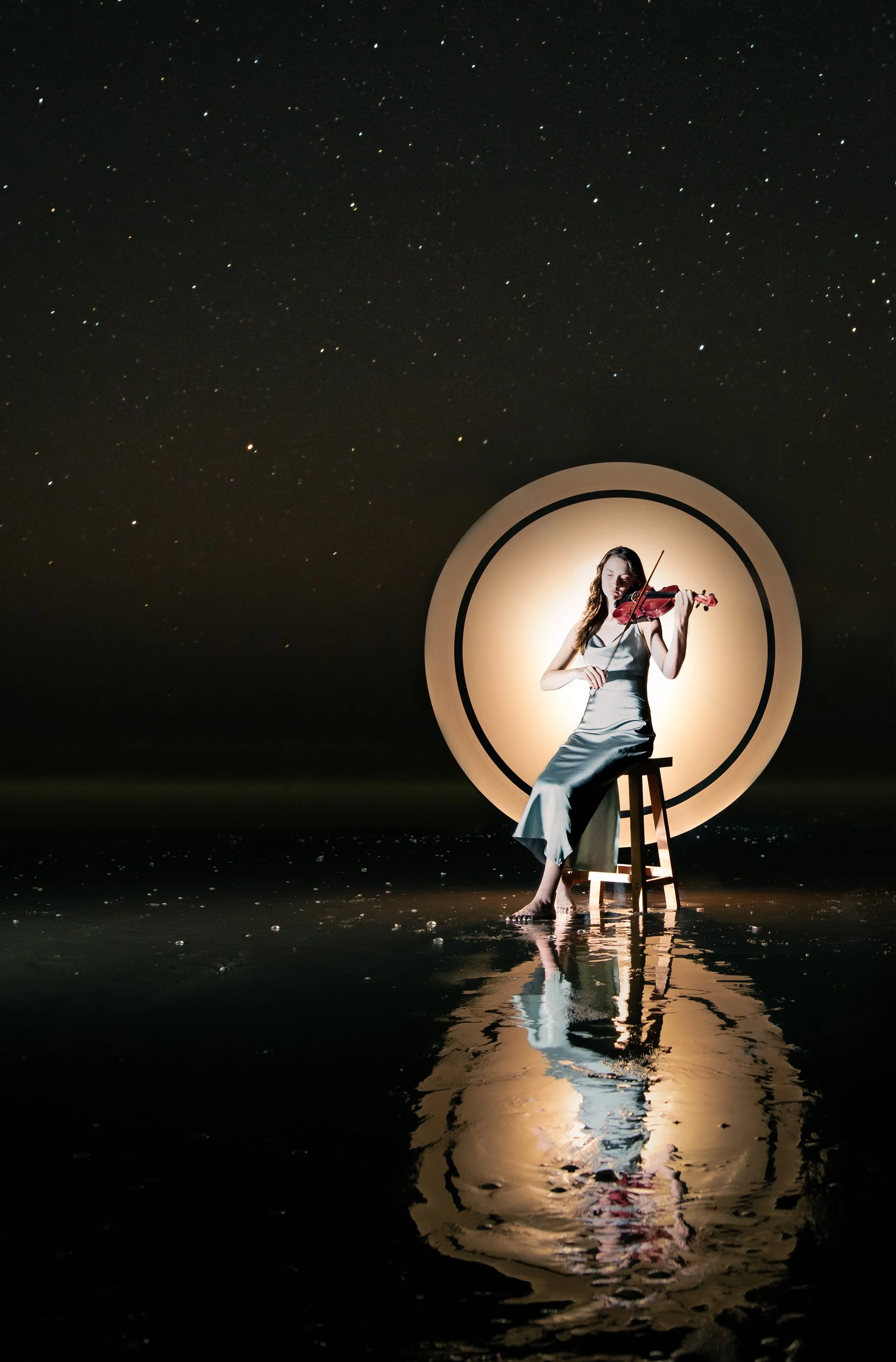 A woman in a gray dress is sitting on a stool, playing the violin, illuminated by a circular light, with a starry night sky in the background and reflection on water.