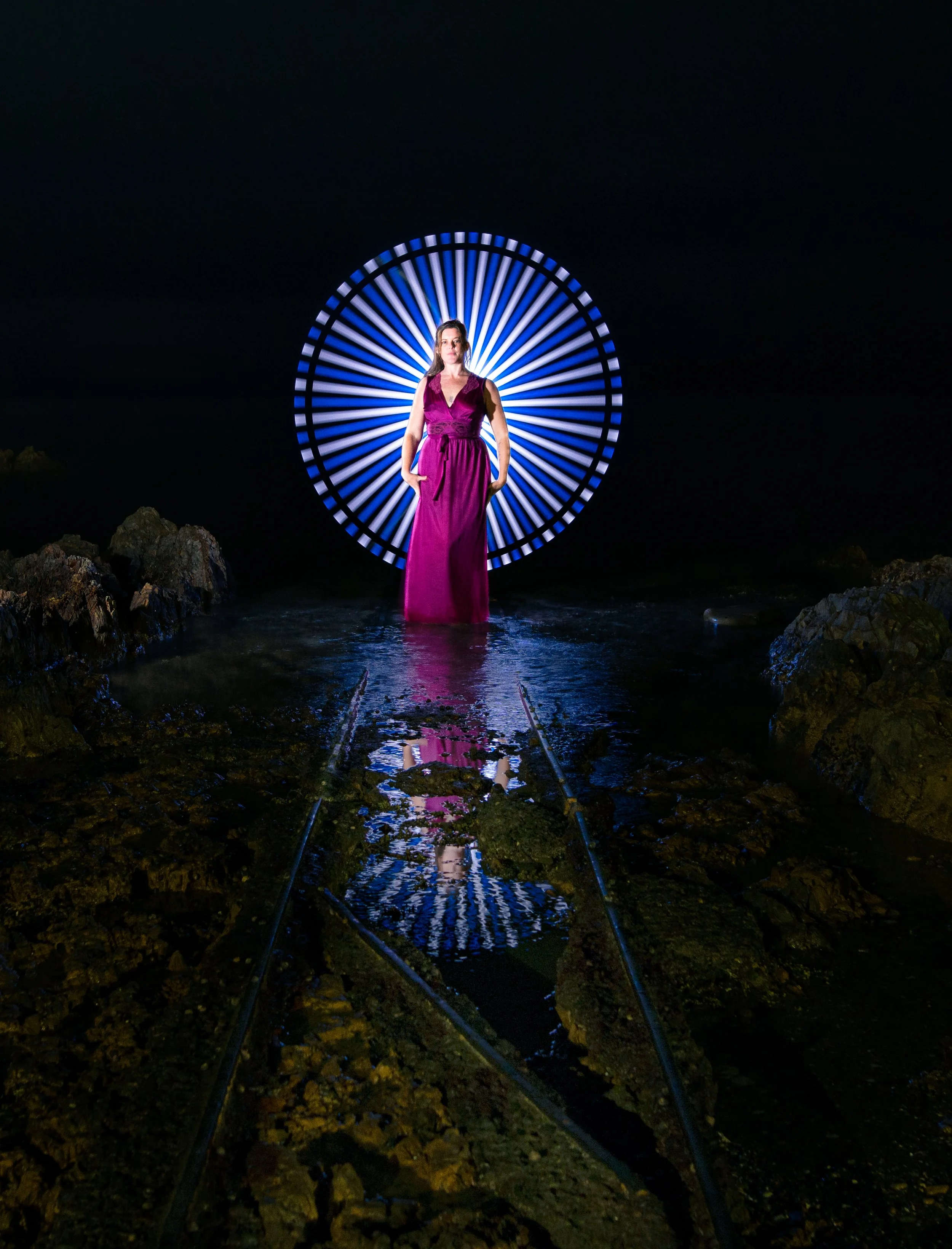 A woman in a purple dress stands on a reflective wet pathway in front of a circular illuminated light installation resembling a ferris wheel against a dark night sky. Rocks are on either side of the pathway in a natural setting.