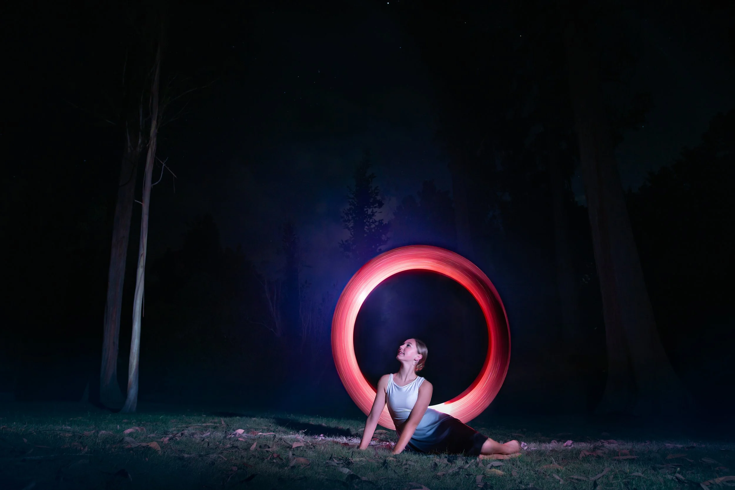 A woman with a braided hairstyle wearing a white tank top and black pants, sitting on the grass at night near tall trees. She is looking up at the sky with stars and a faint moon. A red light trail forms a large circle behind her, creating a glowing 