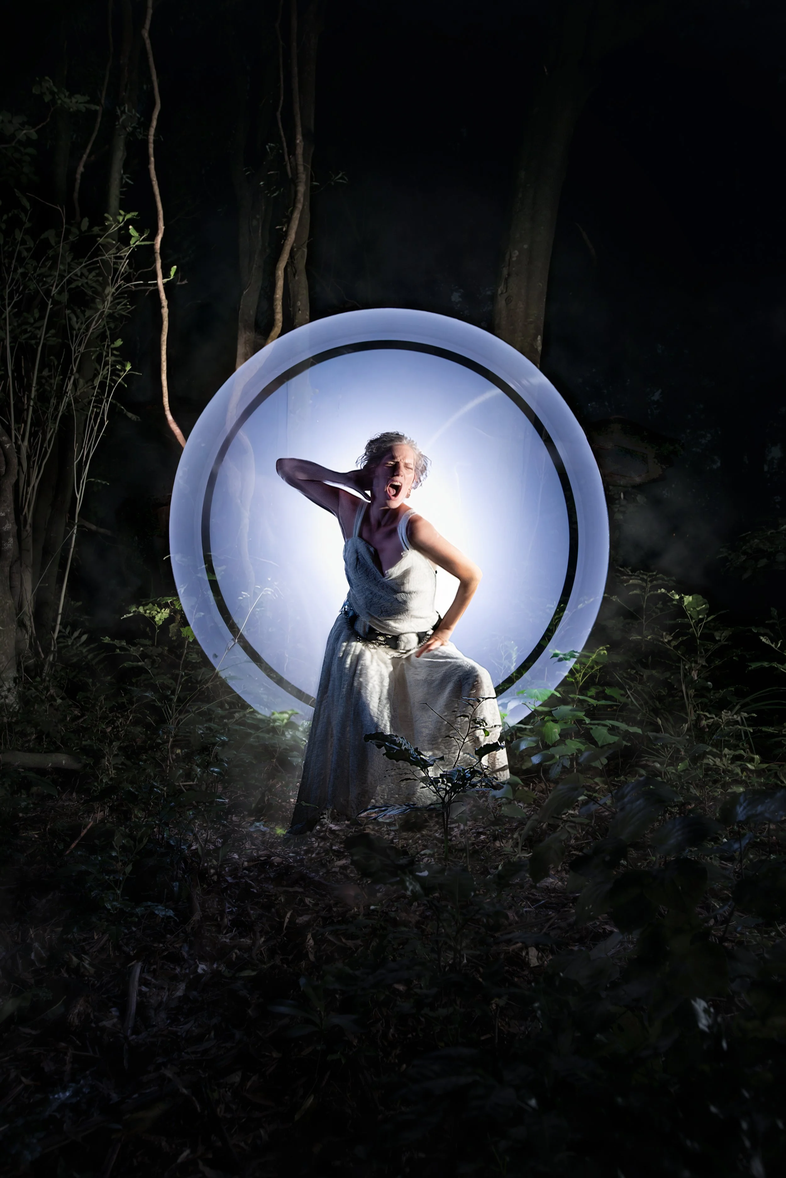 A woman dressed in a long, flowing dress appears to be singing or shouting with her eyes closed and mouth open, standing inside a glowing, circular mirror in the middle of a dark, forested area at night.