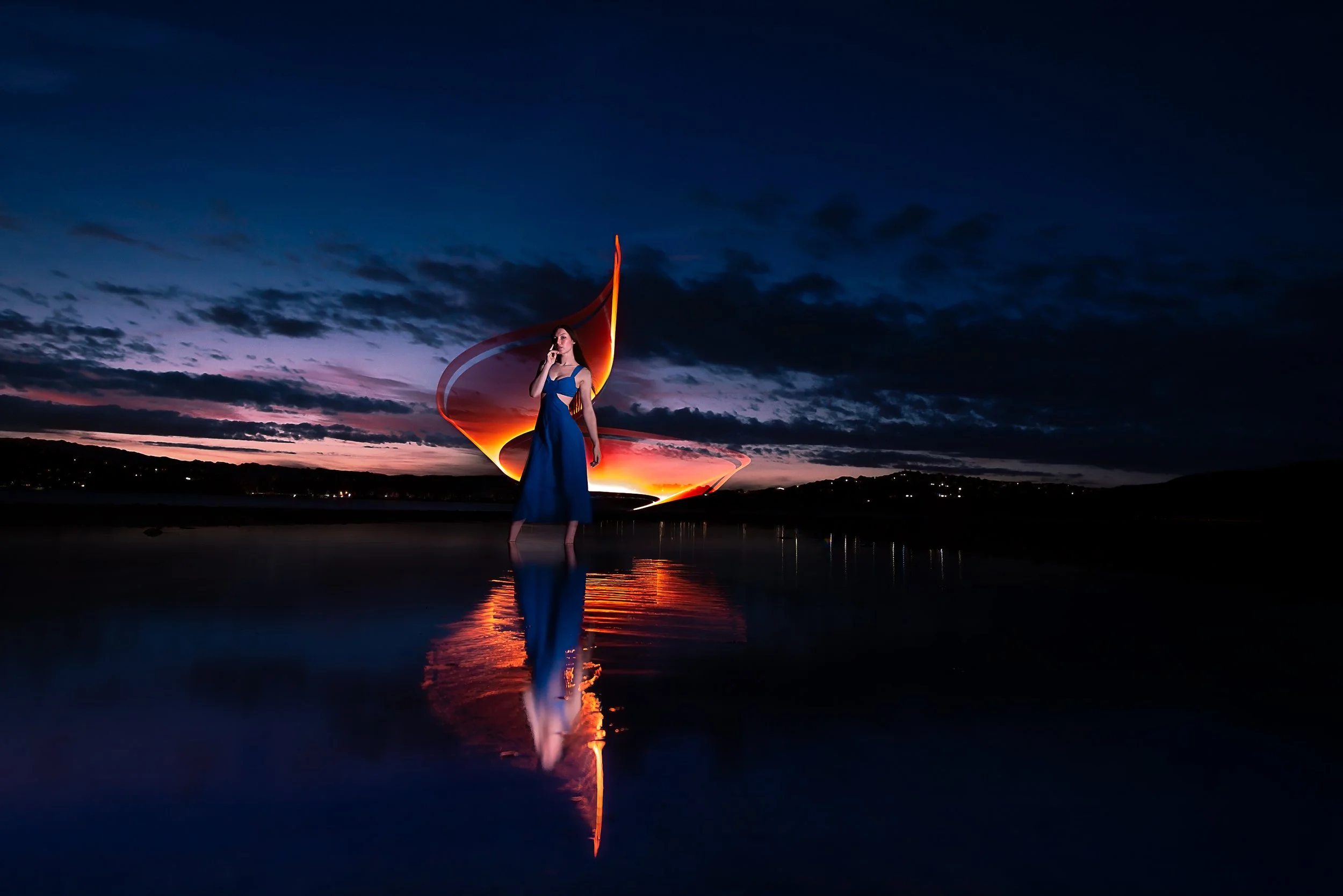 A woman in a blue dress standing near water at sunset with colorful light trails behind her and her reflection visible in the water.