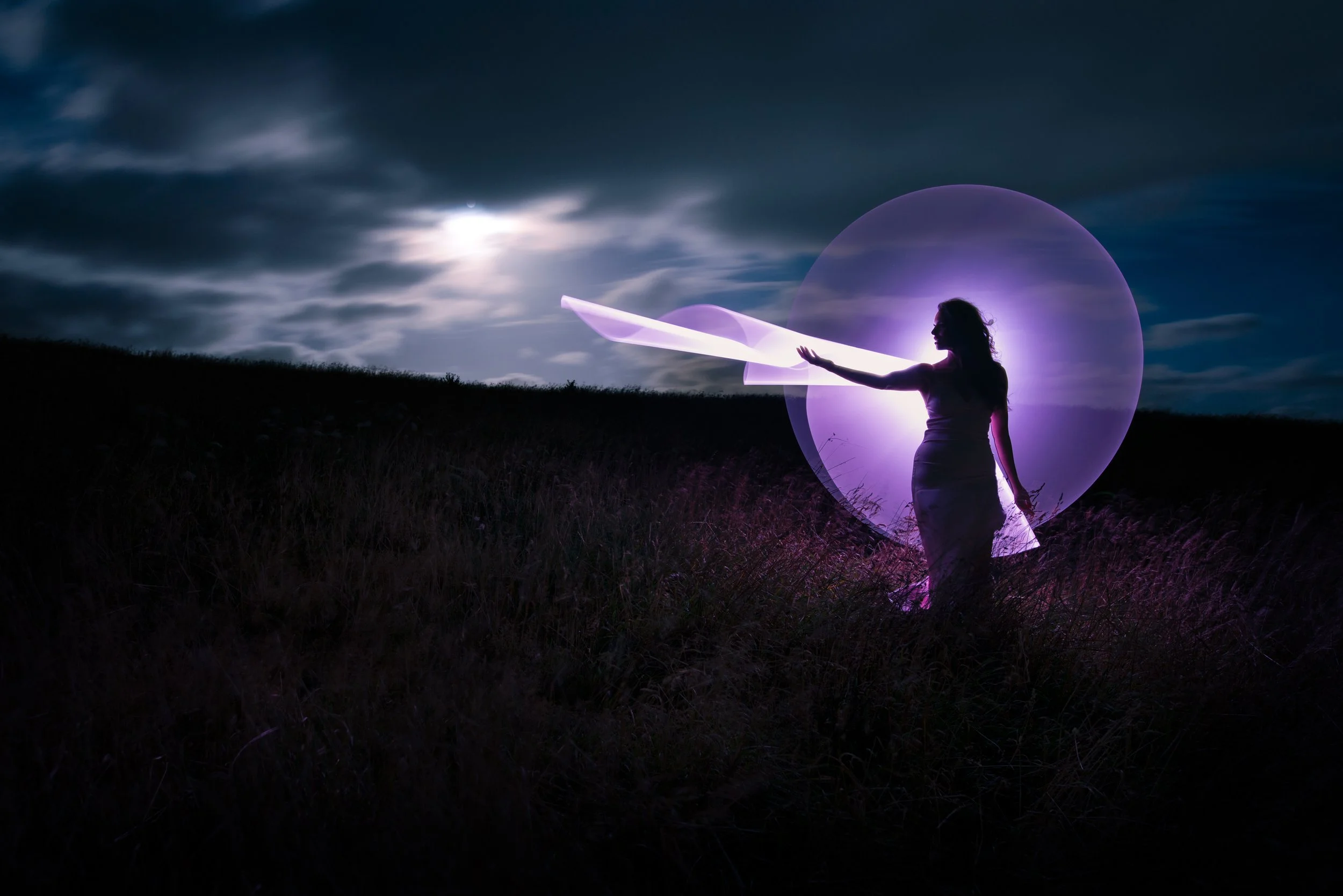 A woman dressed in white standing in a grassy field at night, with glowing purple light swirling around her and a large moon behind her under a partly cloudy sky.