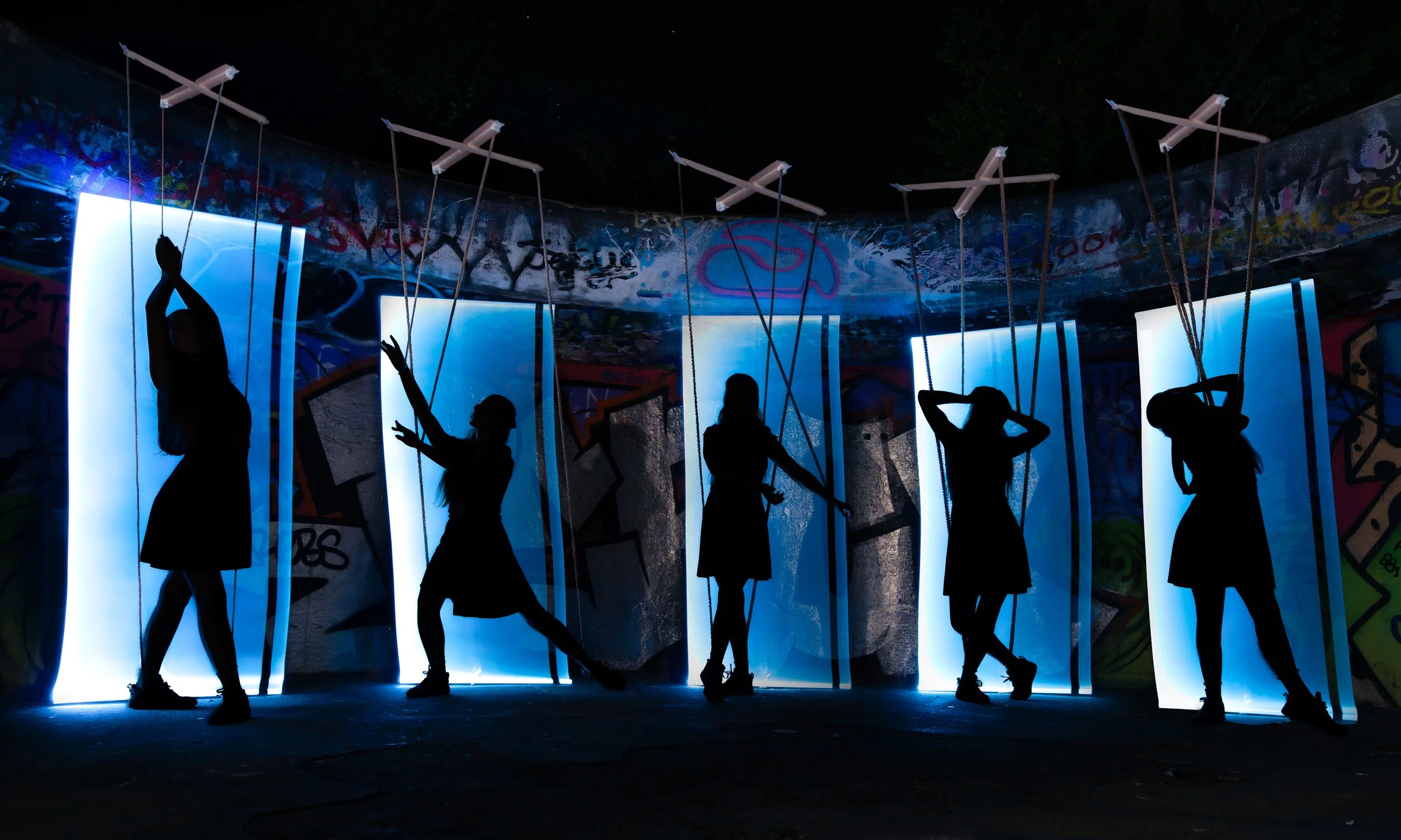 Silhouettes of six women standing in front of illuminated vertical panels and graffiti-covered wall with ceiling of white acoustic panels suspended by ropes, at night.