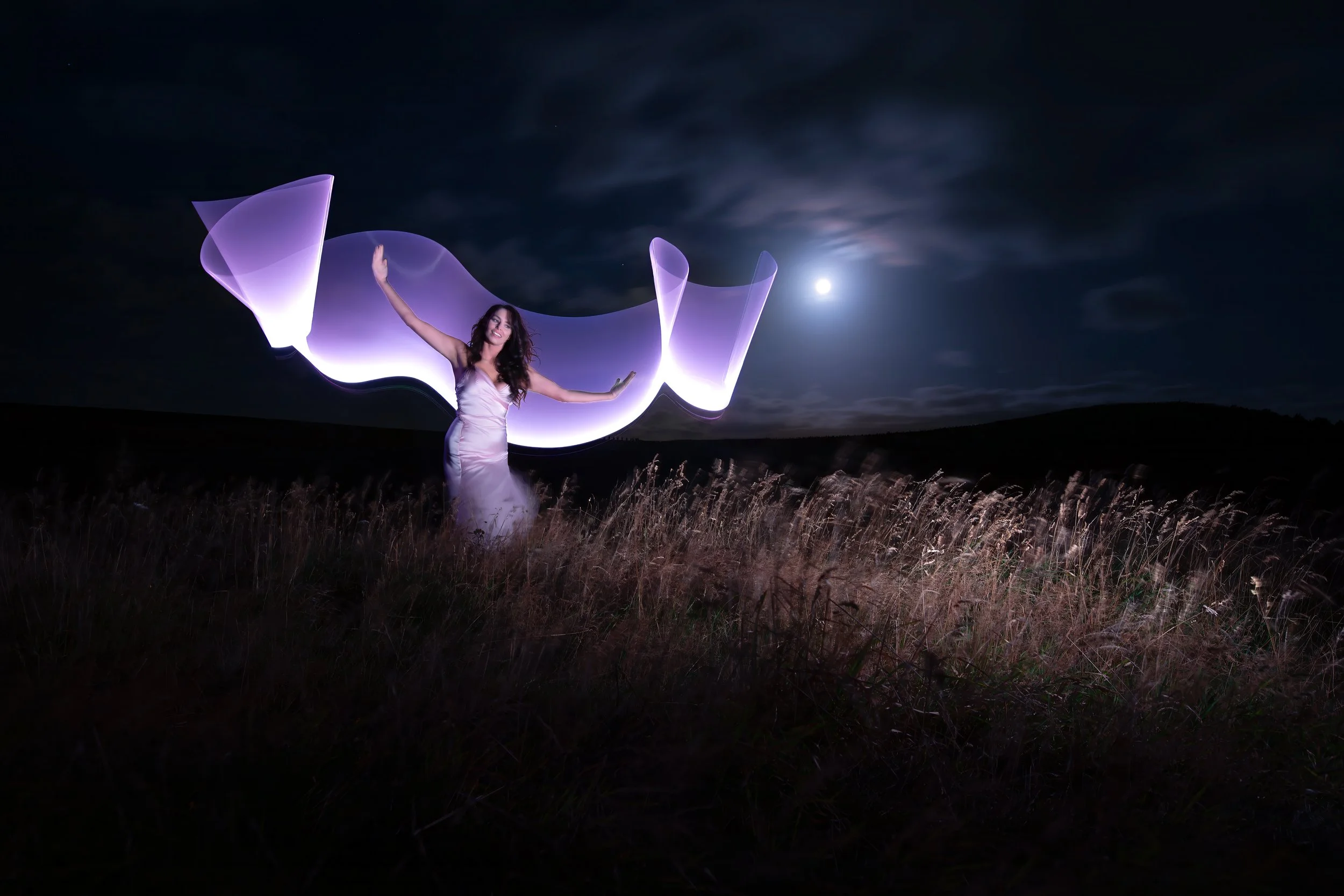 A woman in a white dress standing in a field at night with a light-painting effect around her, under a full moon.