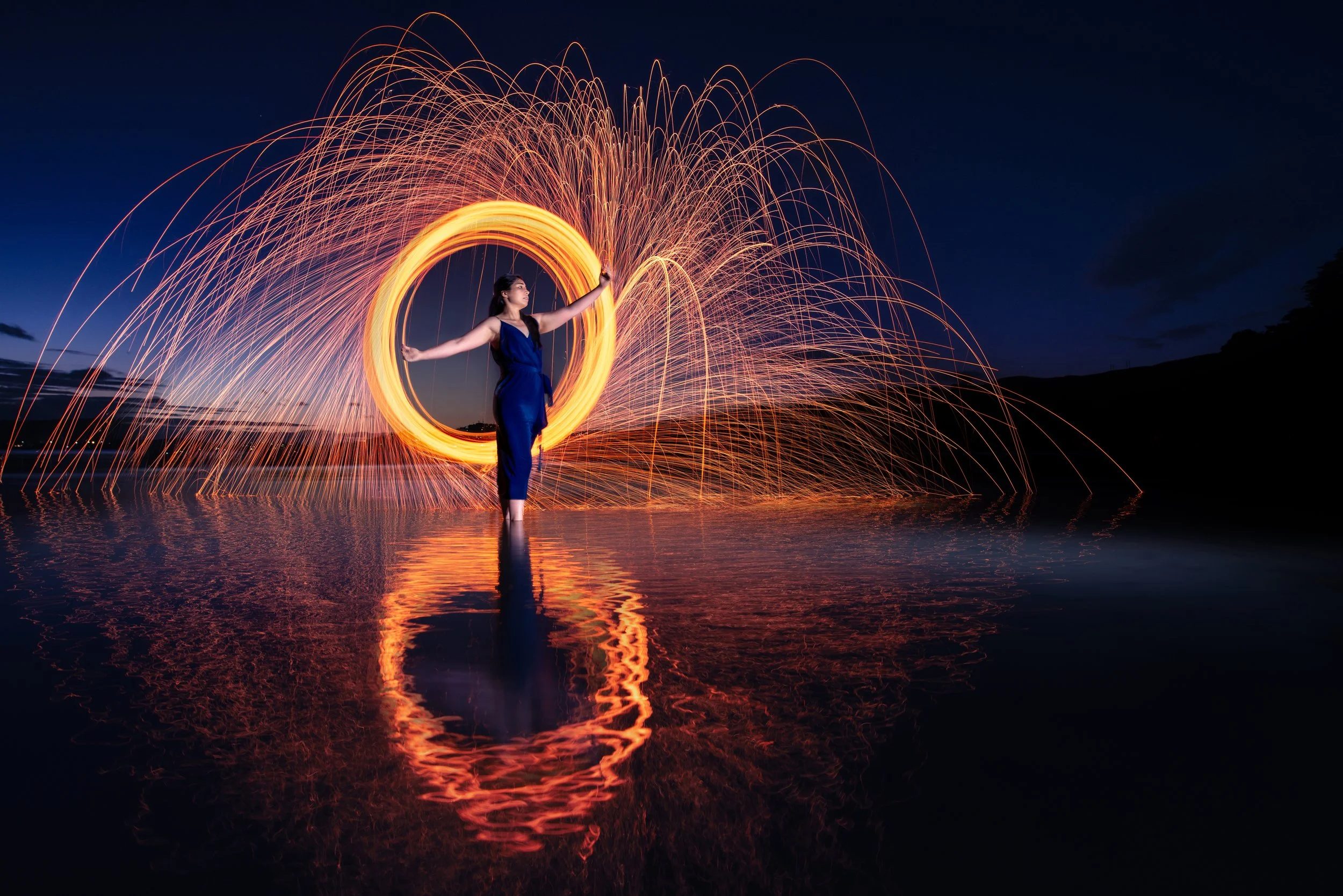 A woman standing in shallow water at night, creating a circle with steel wool spinning, surrounded by sparks flying in arcs, with reflections visible on the water surface.