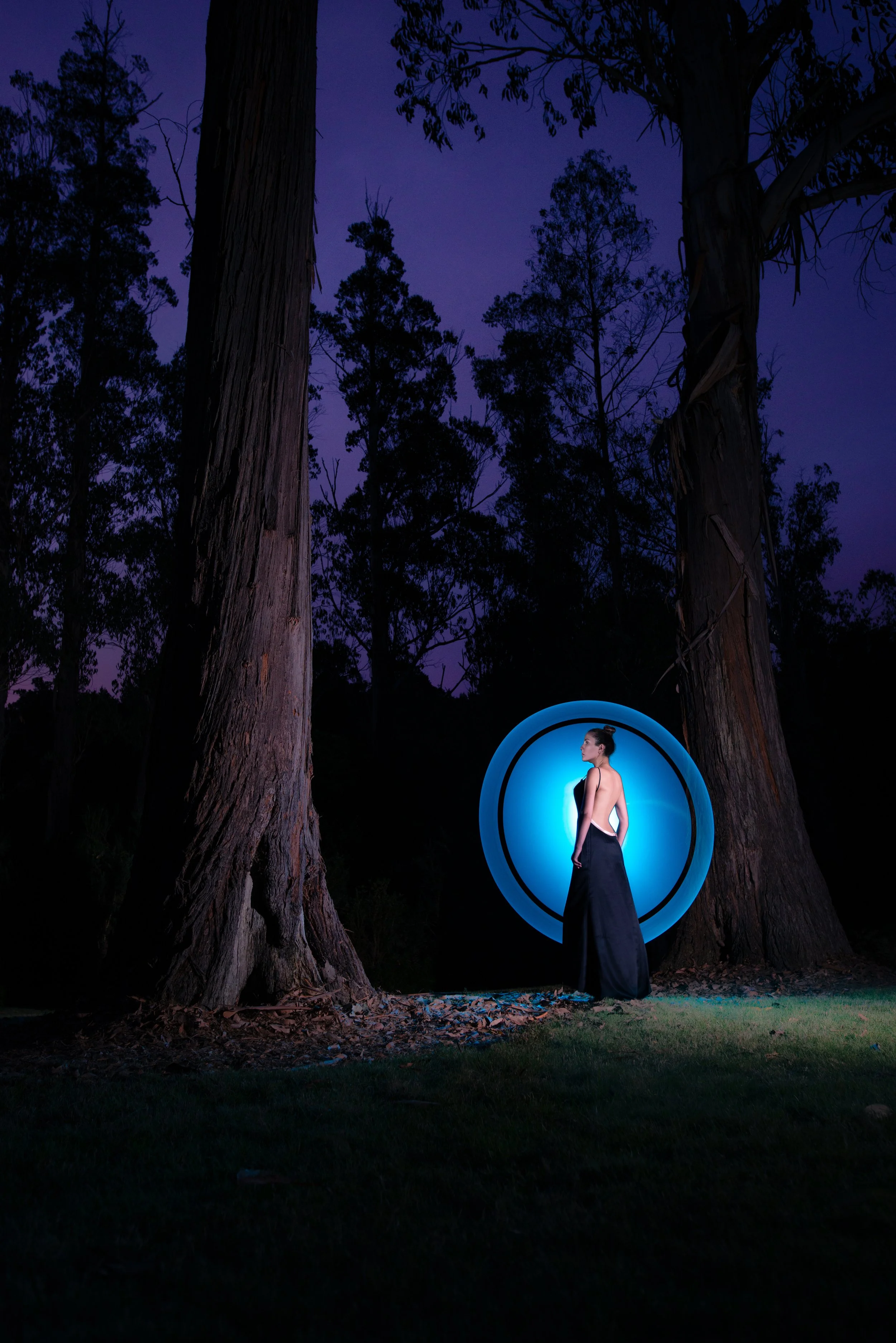 A woman stands outdoors at night inside a blue circular light installation with dark trees in the background, illuminated by the moonlit sky.