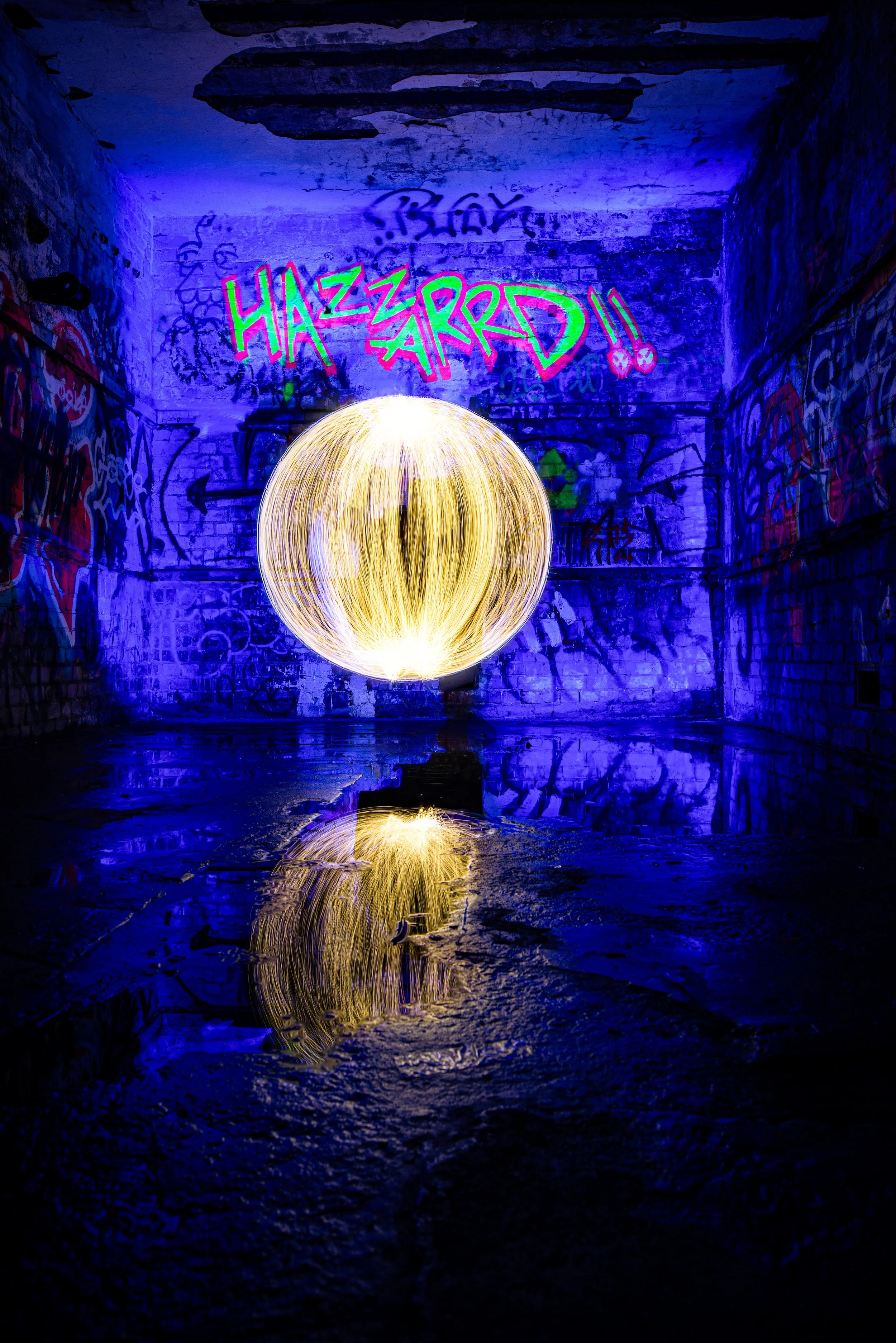 A dark, graffiti-covered underground space illuminated with vibrant blue and purple lighting. In the center, a glowing sphere of yellow light creates a reflective surface on the wet floor, with the reflection of the sphere visible on the ground.