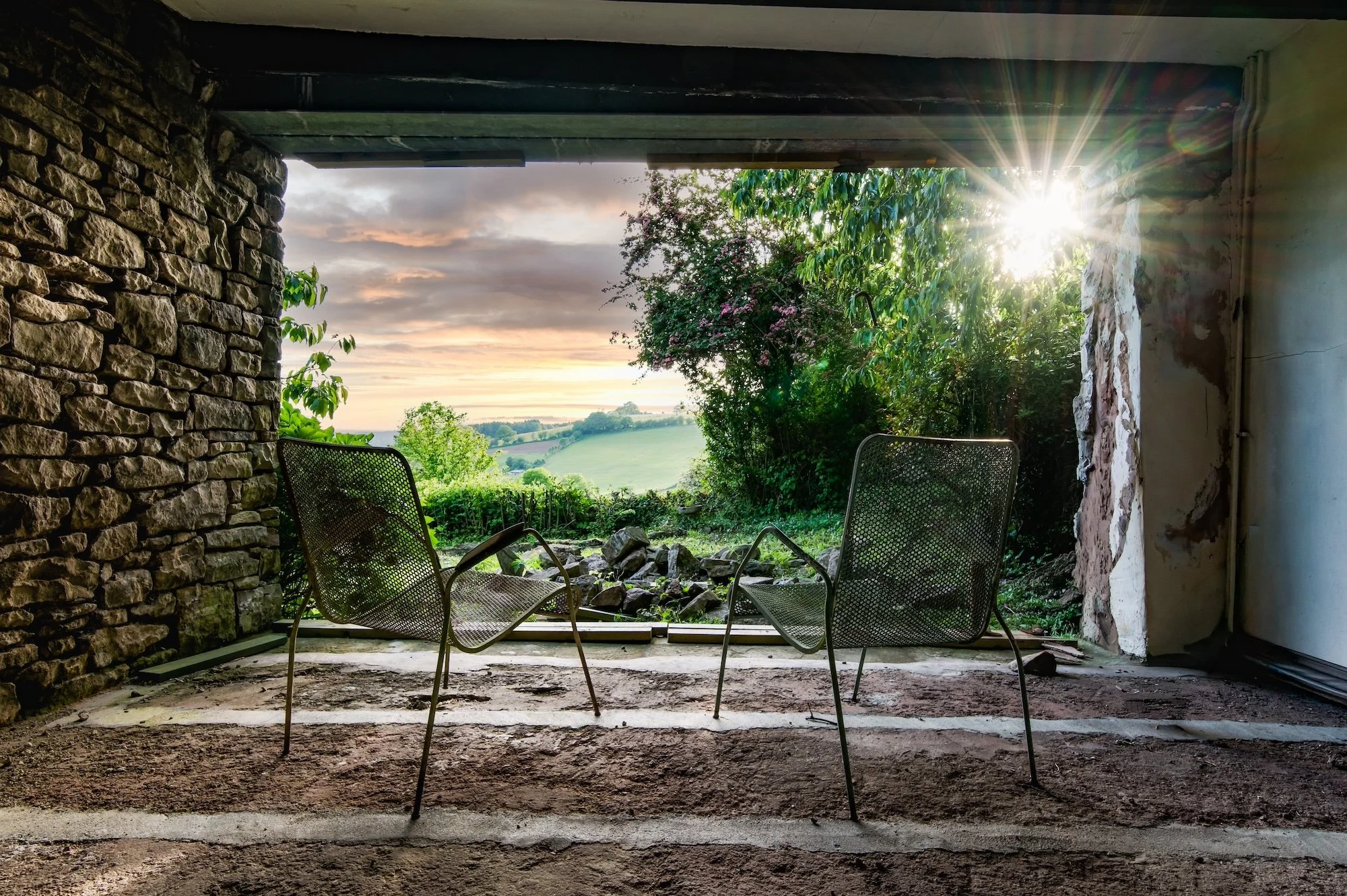 An unfinished abandoned building with exposed brick and concrete walls, three metal chairs with mesh backs sit on the dirt floor, overlooking a lush green landscape with trees, rolling hills, and a sunset sky.