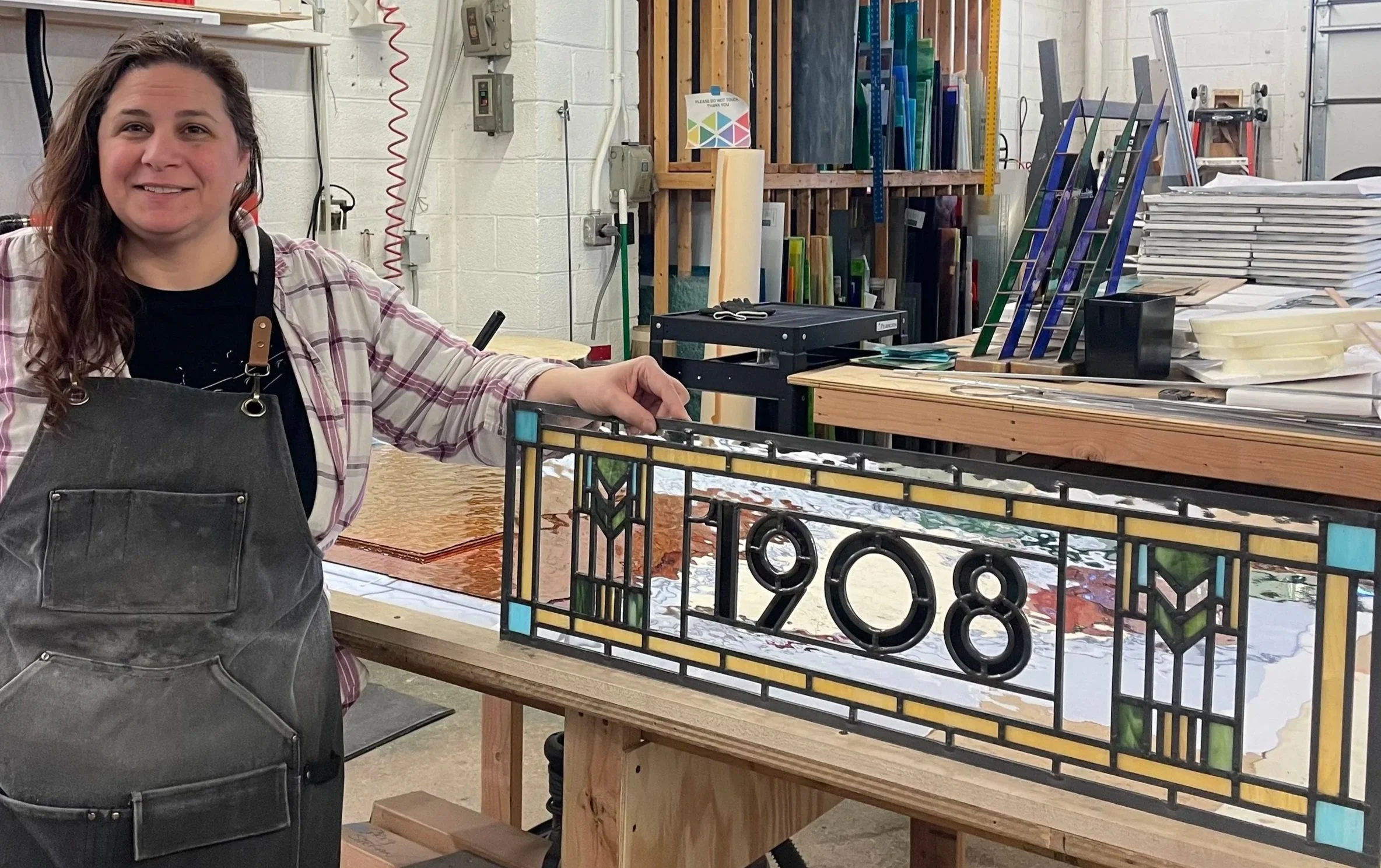 A woman in an art studio displaying a stained glass piece with the number 798 in colorful design. She is smiling, wearing a plaid shirt and dark apron, and leaning on a worktable.
