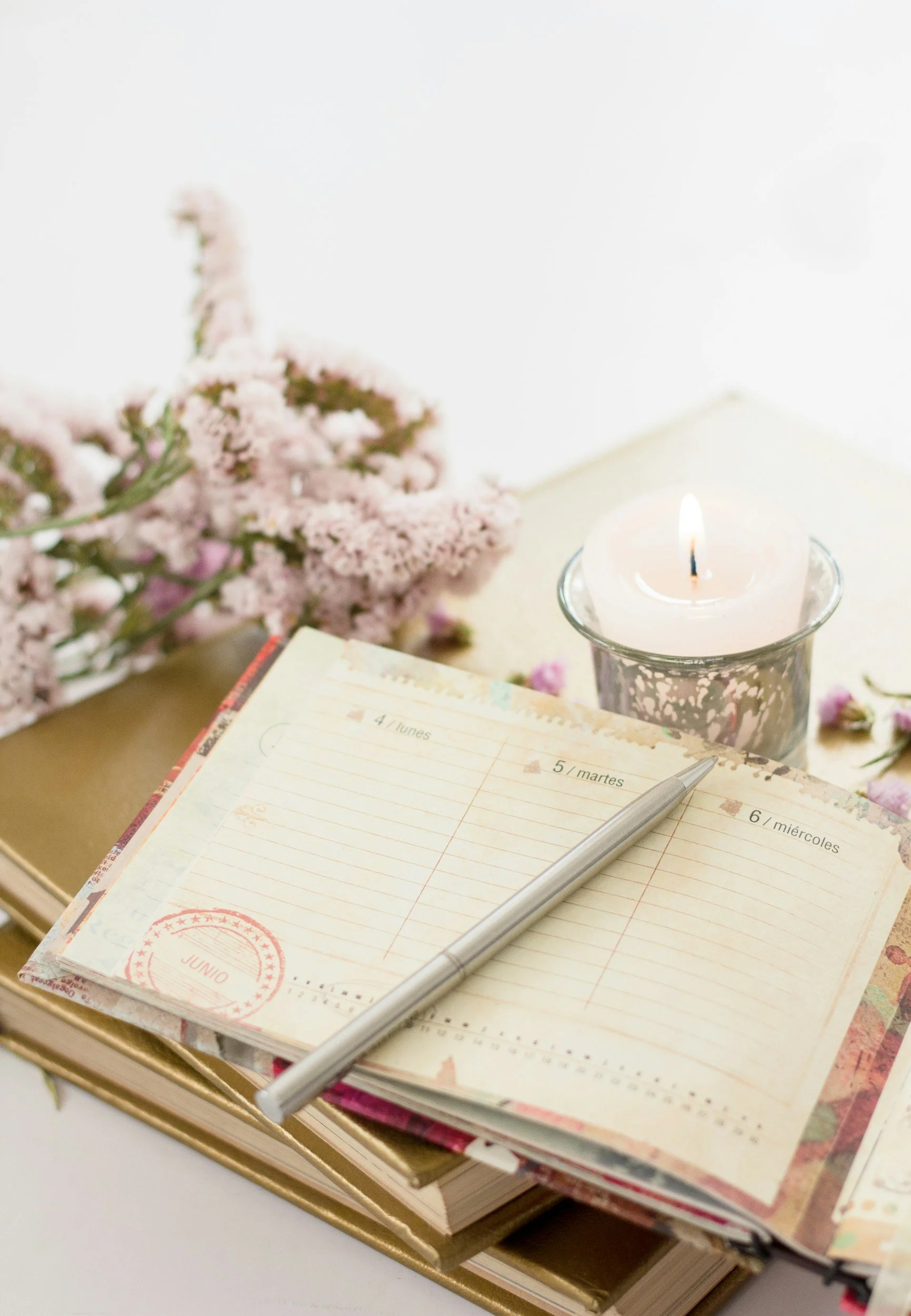 Open planner with handwritten notes, a pen, a lit candle, pink flowers, and a book on a desk.