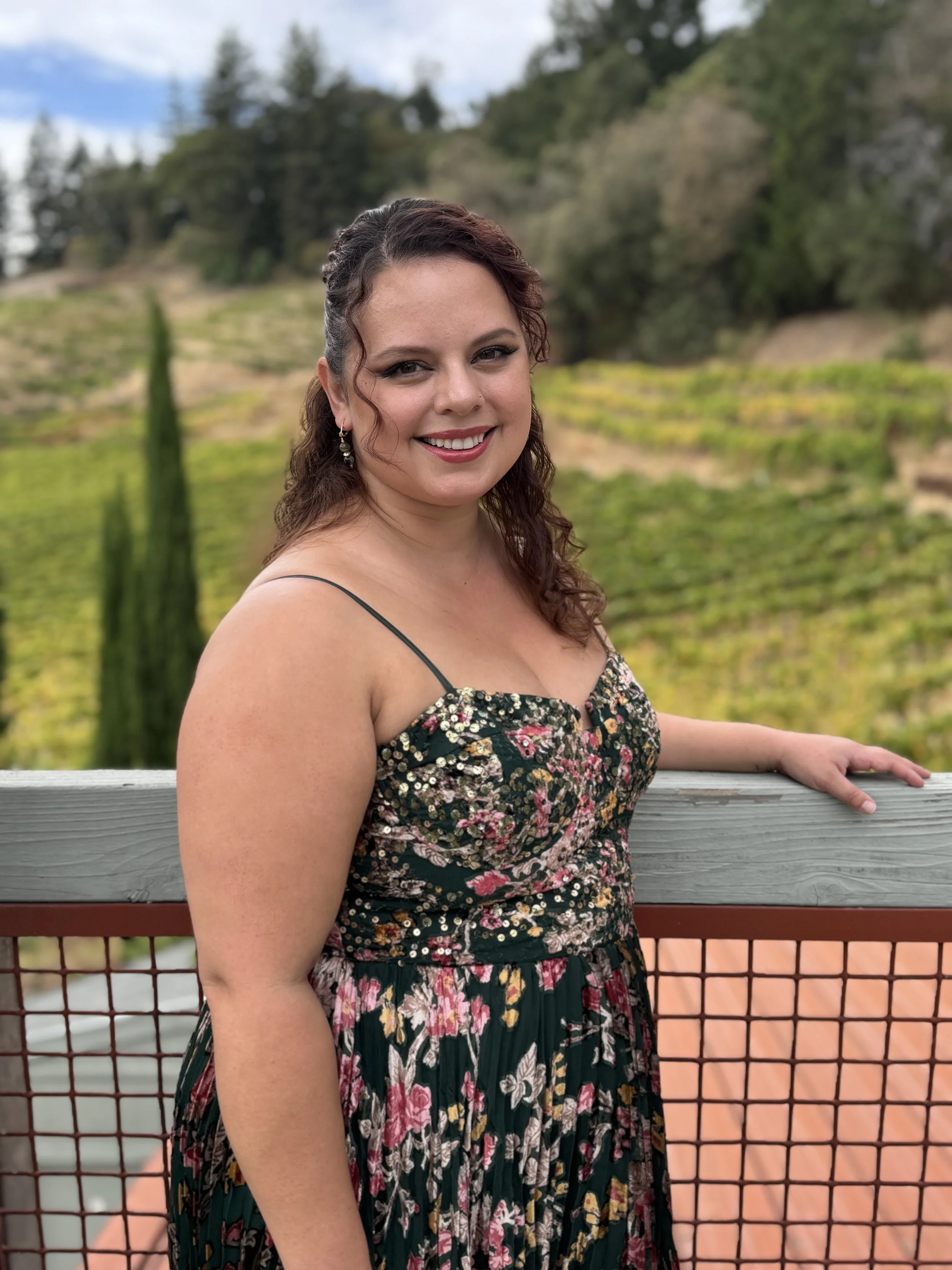 A woman with brown hair and earrings smiling at the camera, standing outdoors by a railing with a scenic landscape of trees, hills, and cloudy sky in the background.