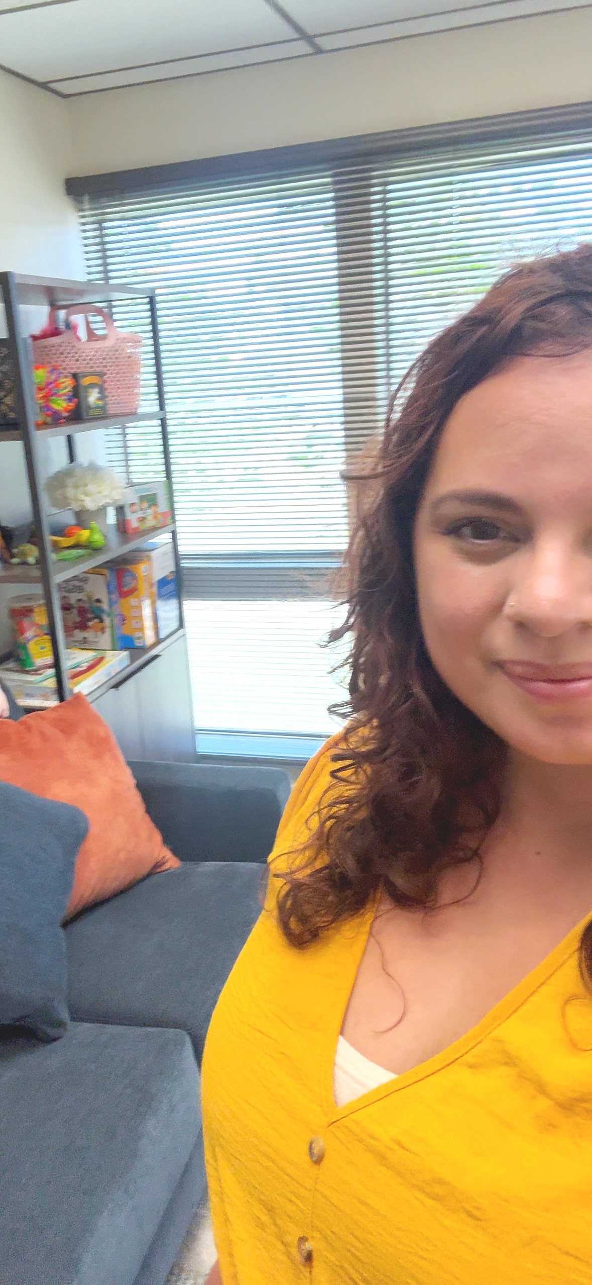 Close-up of a woman with brown curly hair, wearing a yellow top, smiling in a cozy living room with a dark gray sofa, orange throw pillow, bookshelf with toys and decorations, and a window with blinds in the background.