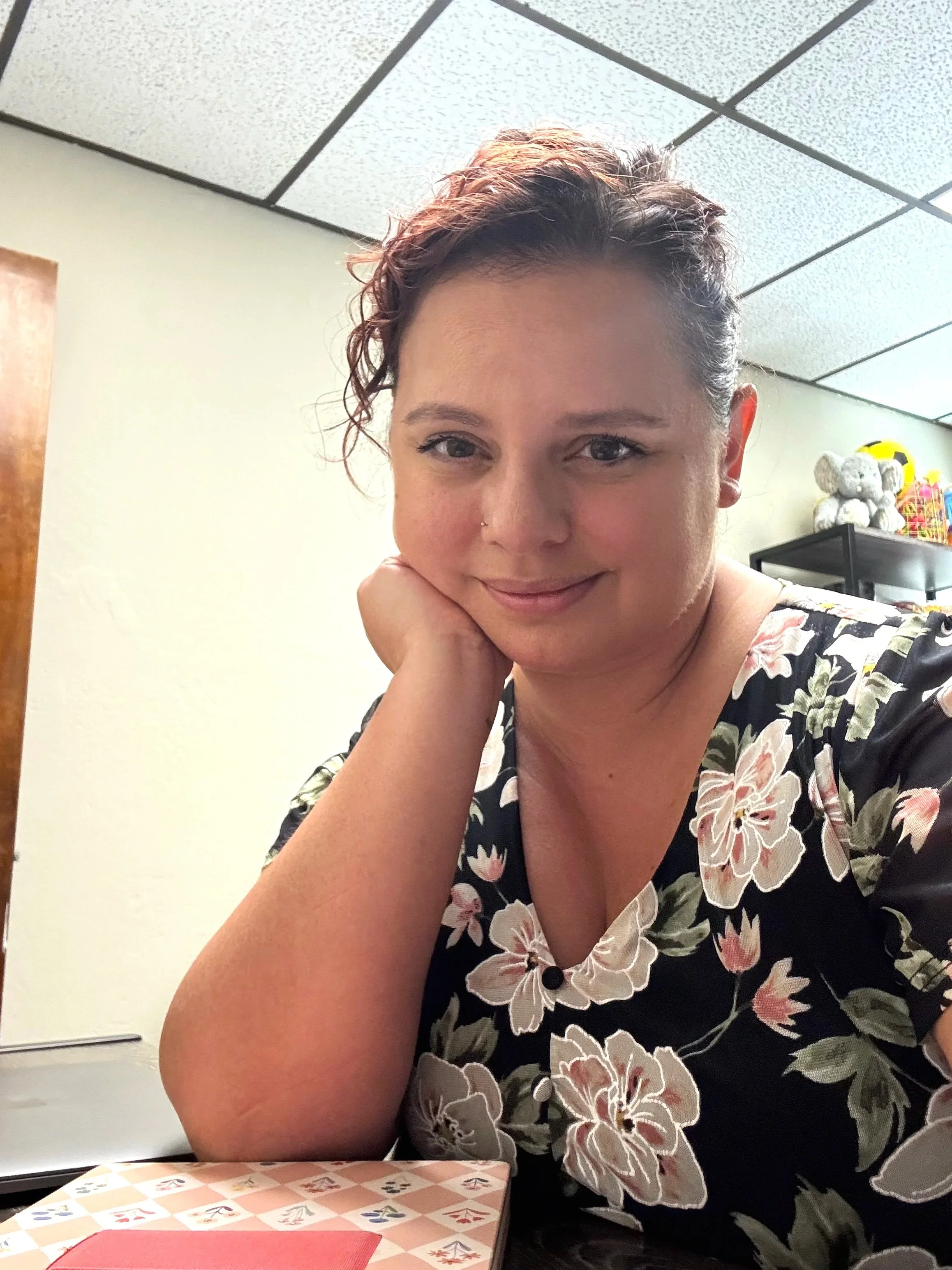 A woman with short, curly hair and a nose piercing sitting at a table in a room with a textured ceiling. She is resting her chin on her hand and smiling at the camera. There are some plush toys and colorful items on a shelf in the background.