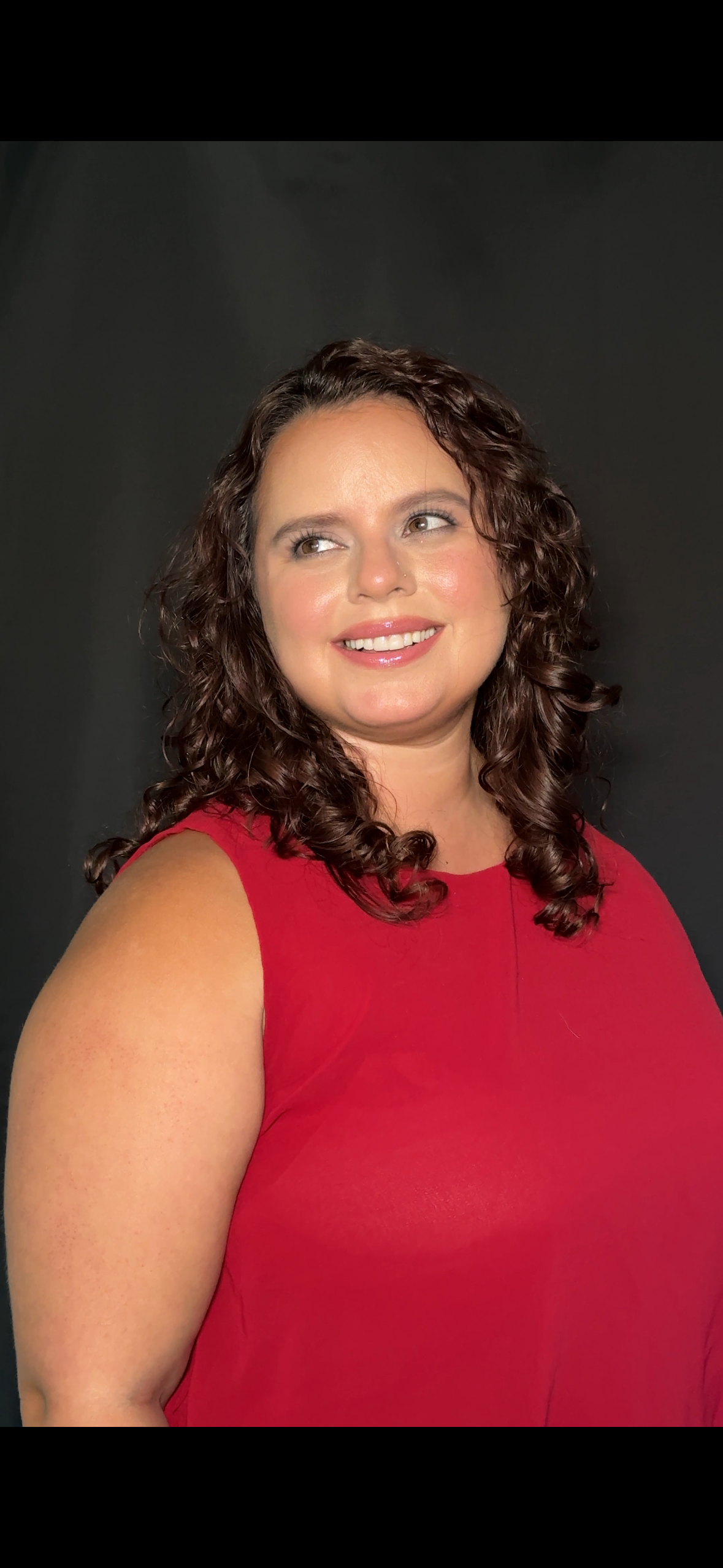 Portrait of a woman with curly brown hair wearing a red sleeveless top, smiling and looking slightly to the side against a dark background.