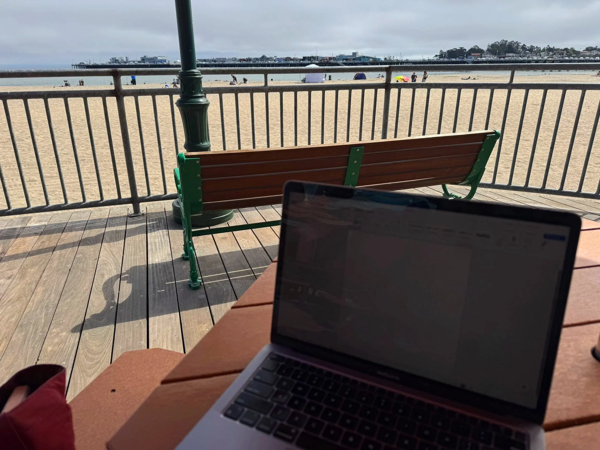A person working on a laptop at a beachside wooden table, with a view of the sandy beach, railing, and ocean in the background. There are people on the beach and a pier visible in the distance.