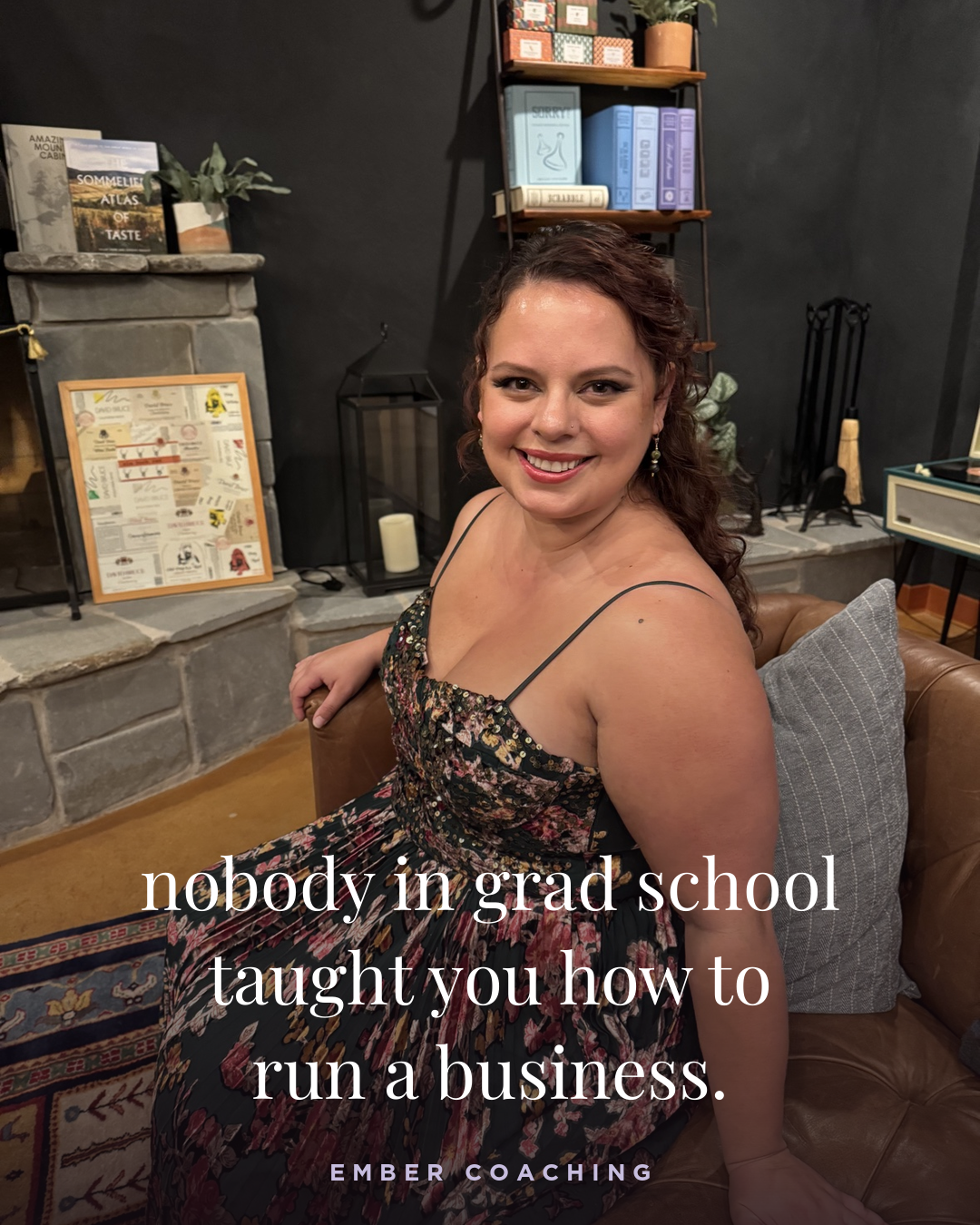 A smiling woman in a floral dress sitting on a leather couch in a cozy room with a stone fireplace, books, and decorative items.