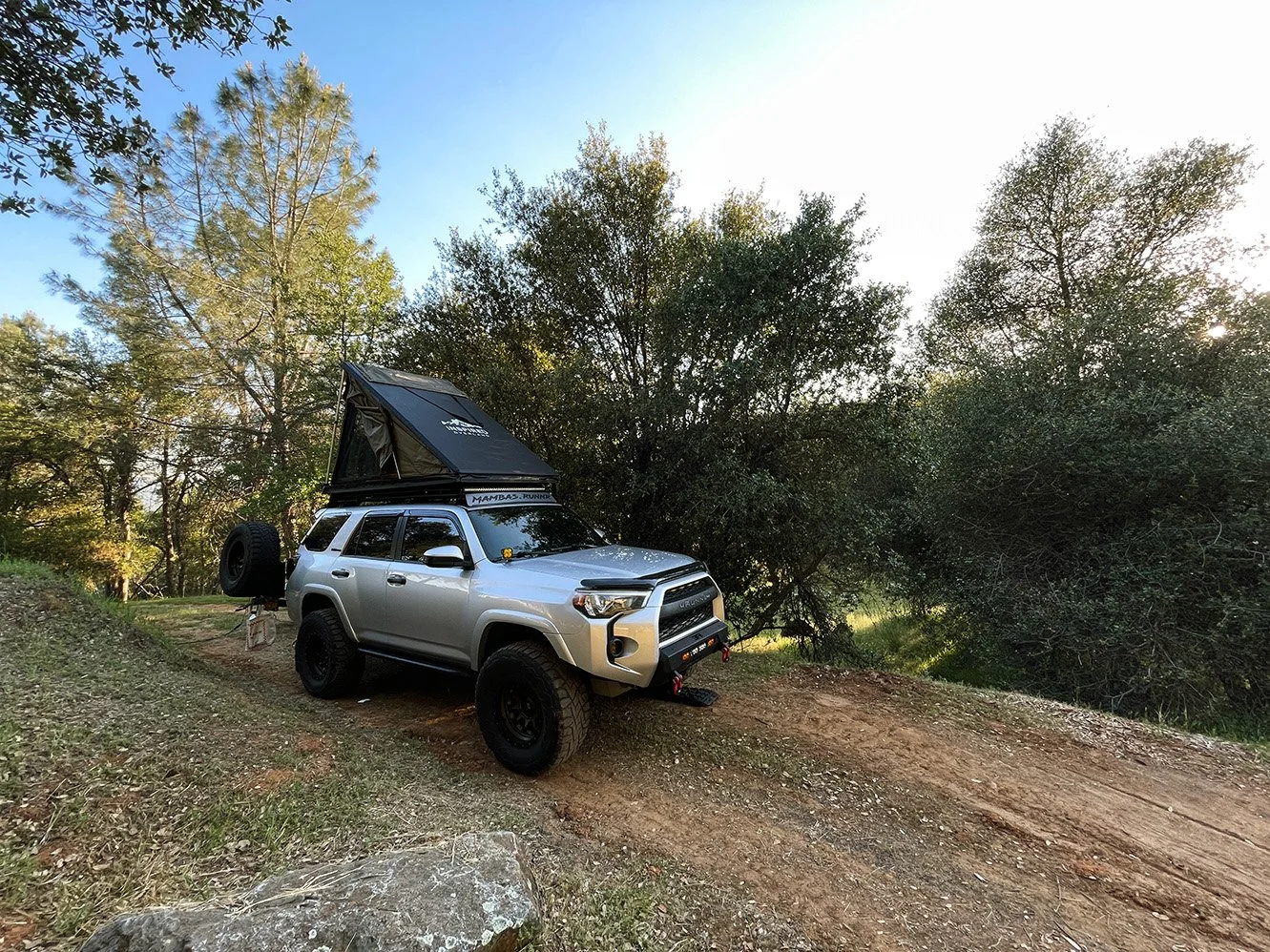 Silver off-road vehicle with a rooftop tent, parked on a dirt trail in a wooded area during daytime.