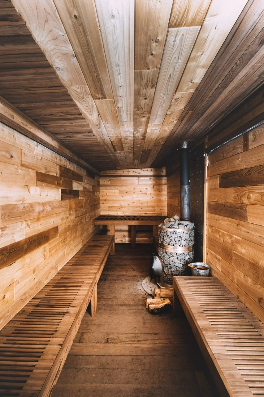 Inside a wood-paneled sauna with wooden benches on both sides and a wood-burning stove with rocks in the corner.