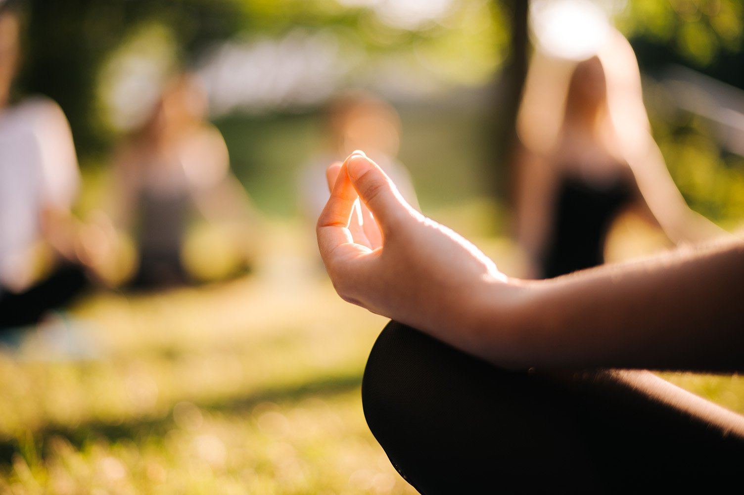 A person practicing yoga outdoors, sitting cross-legged and making a meditation mudra with their hand, with others in the background blurred.