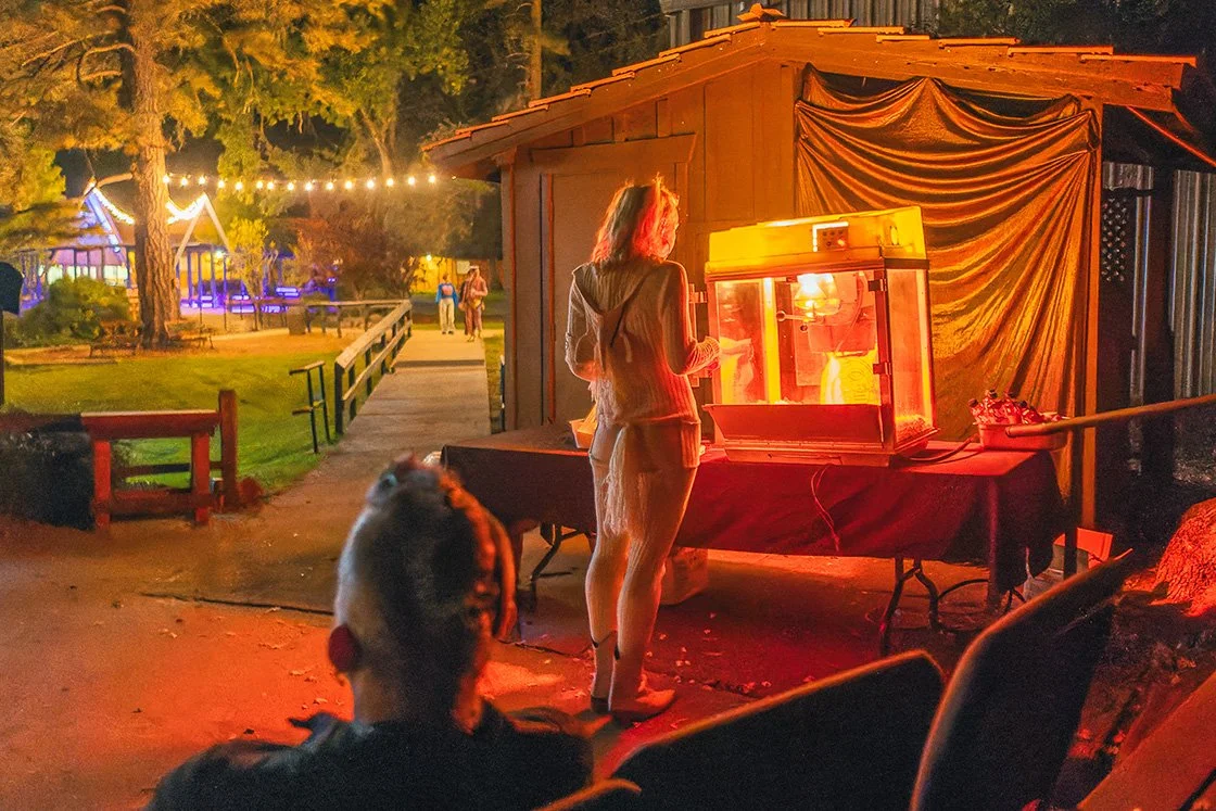 A woman stands at an outdoor carnival game booth at night, illuminated by the red glow of a ticket dispenser, with a dog sitting nearby in foreground, and a playground with string lights in the background.